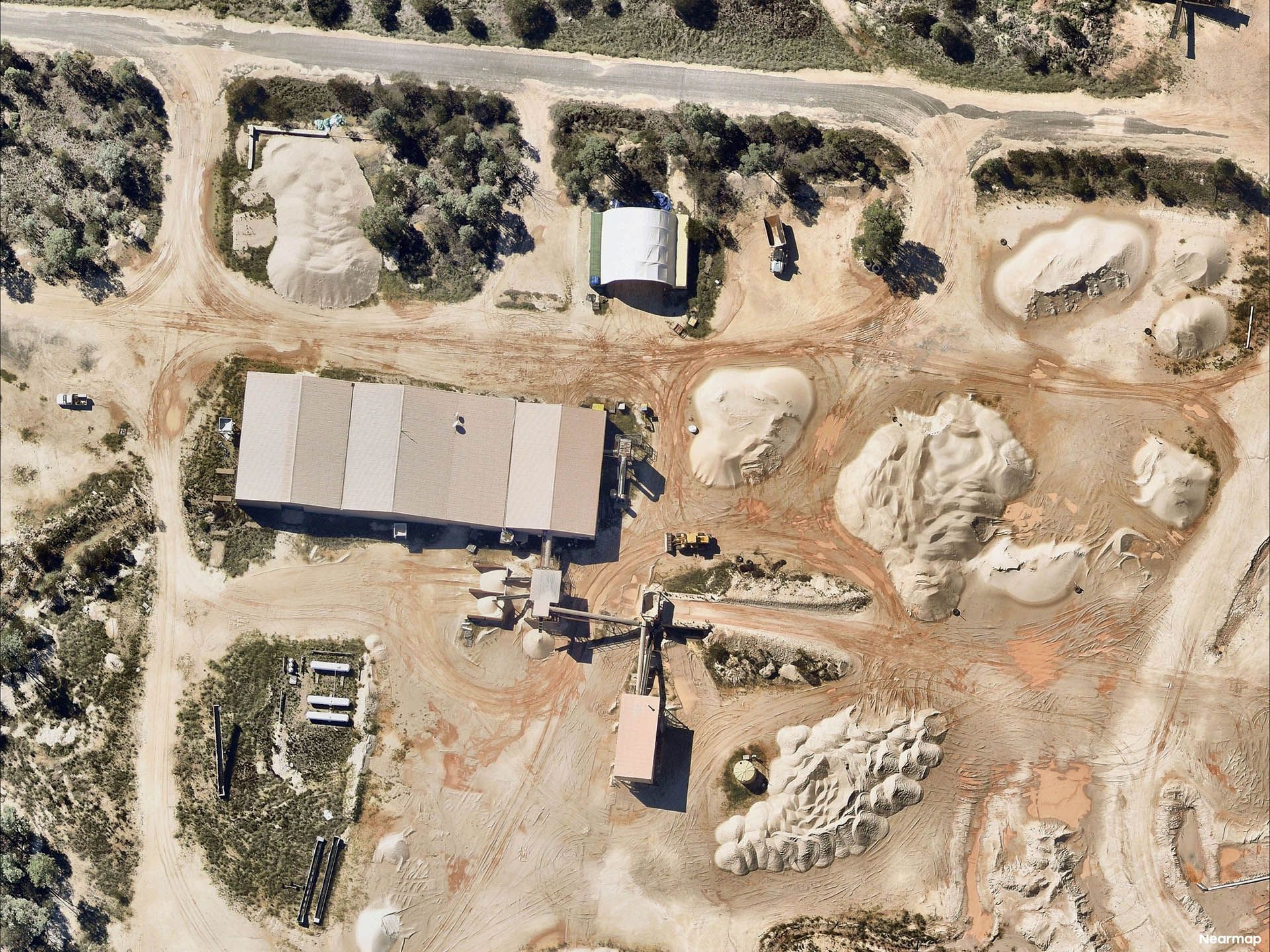 Aerial view of a dusty quarry with a warehouse, vehicles, dirt mounds, and surrounding trees and road