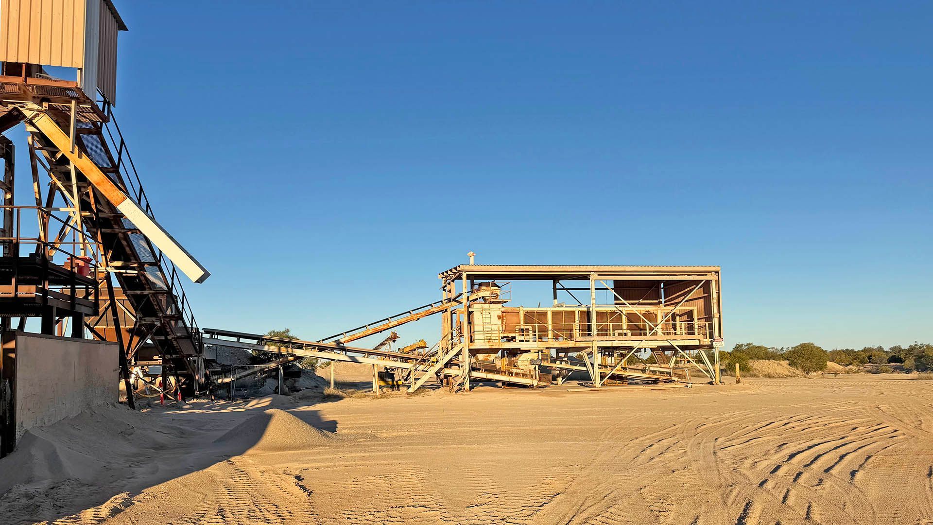 Desert industrial site with metal conveyors, a skeletal building frame, and sandy ground under a clear blue sky