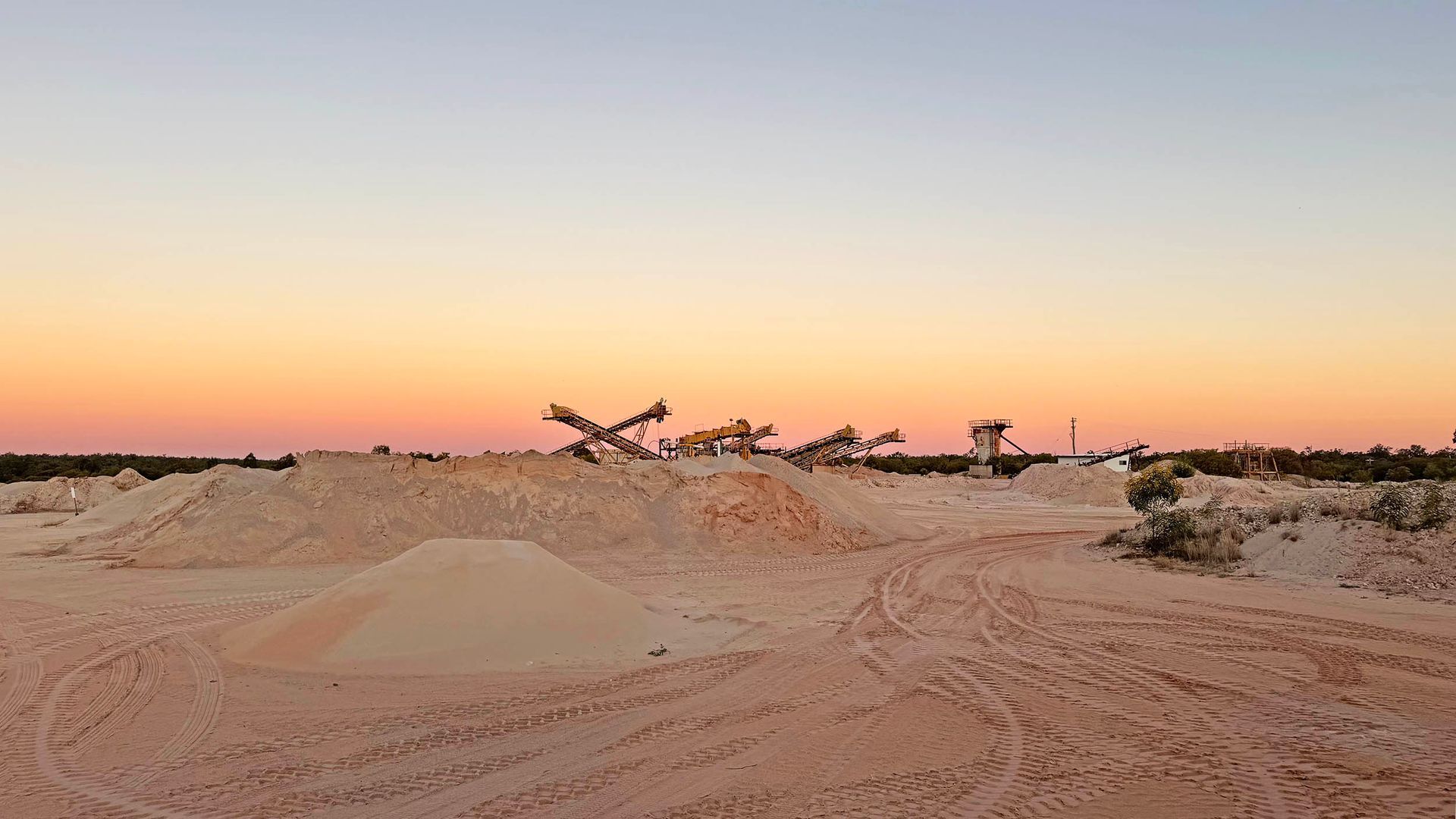 Sandy desert at sunset with low dunes and sparse shrubs under a pink-orange sky