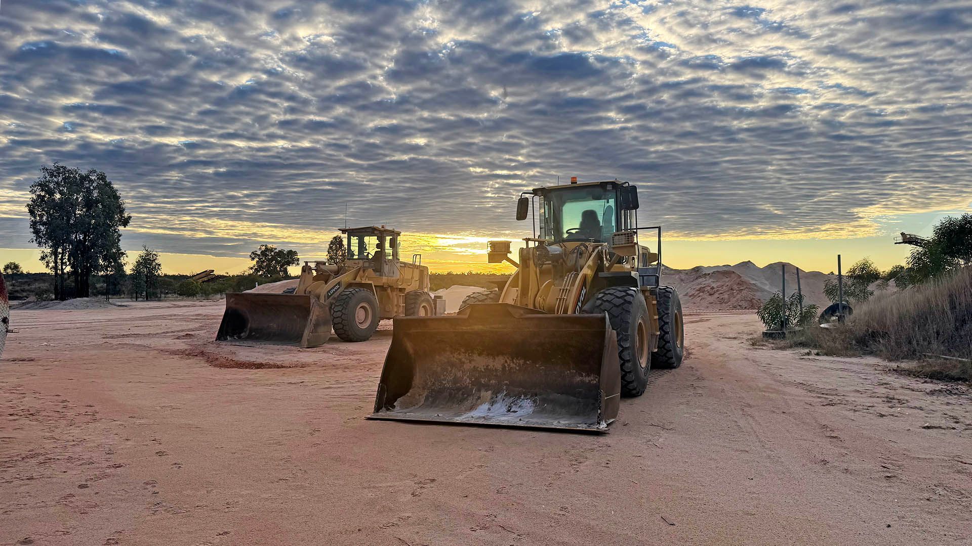 Two bulldozers on a dirt road at sunset under a cloudy sky