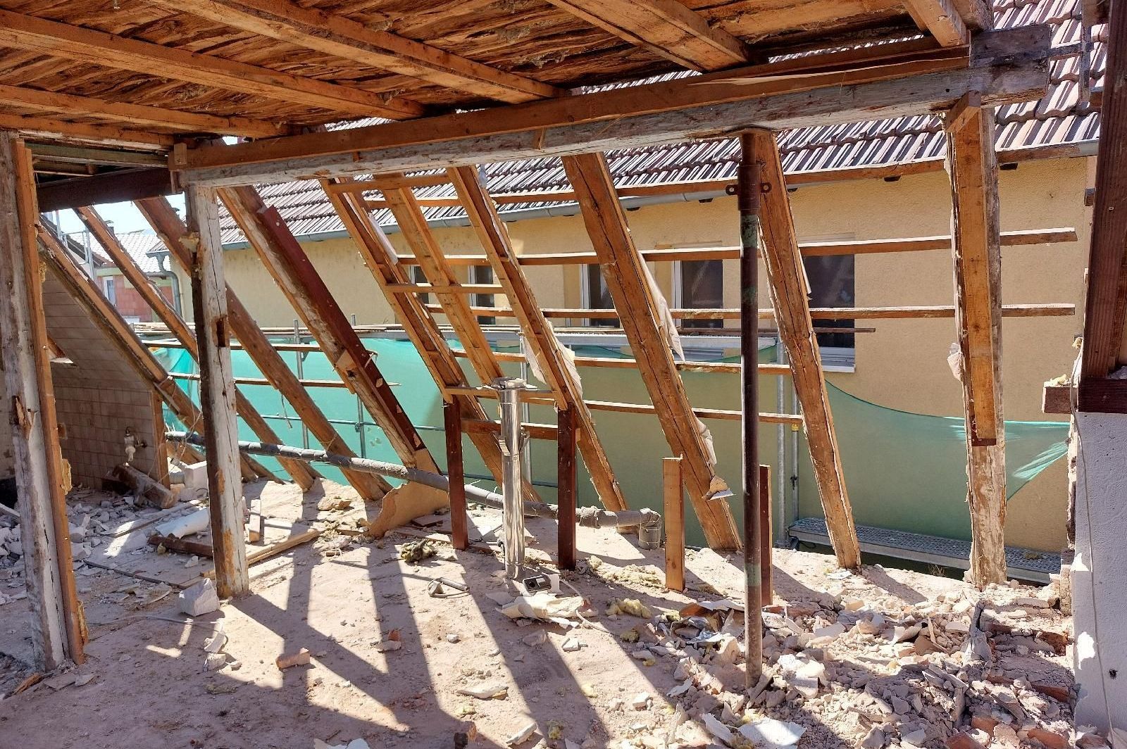 Ein halb abgerissener Dachstuhl. Auf dem Boden liegt Schutt und durch die freiliegenden Balken kann man das Nachbarhaus sehen