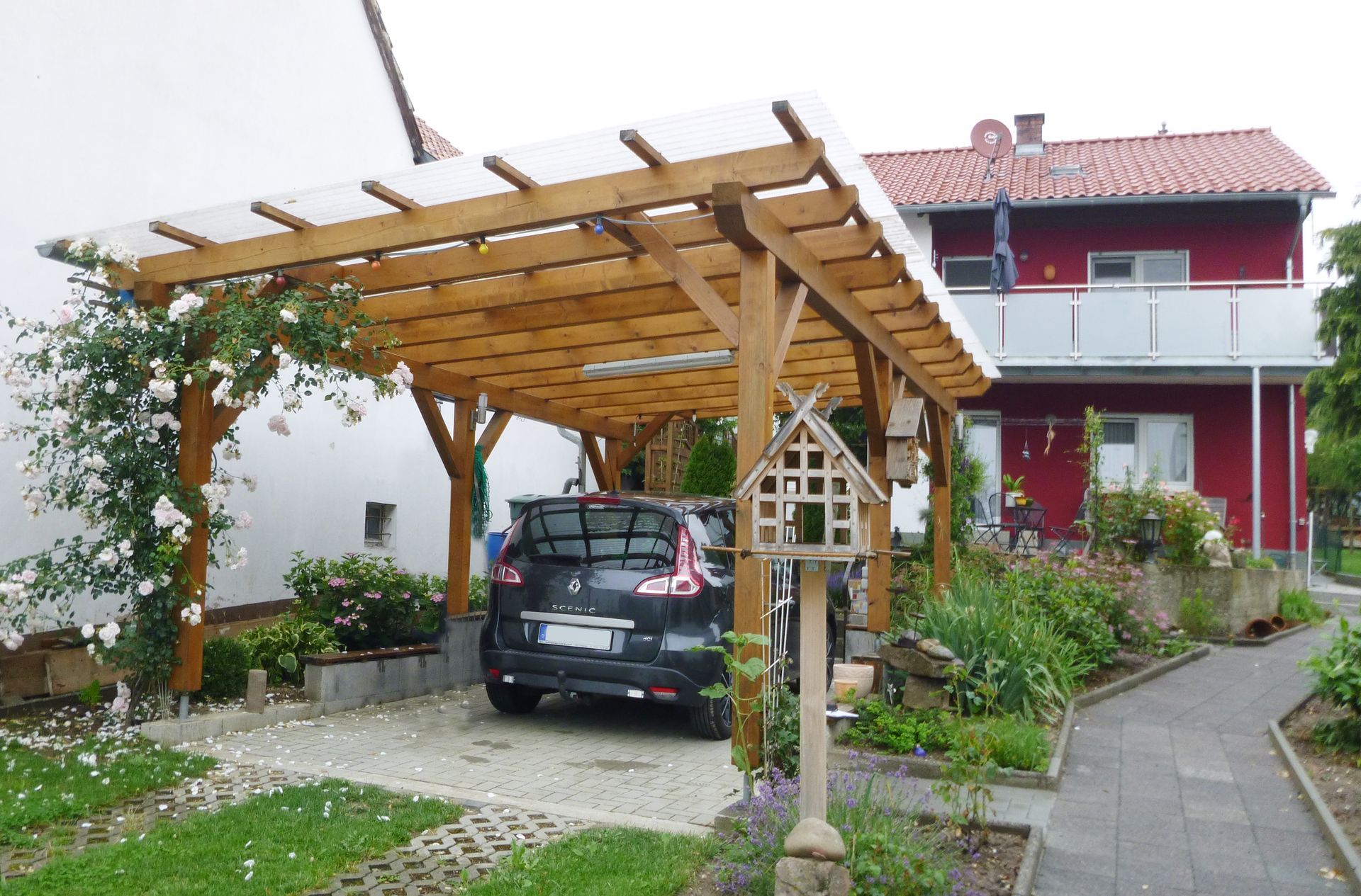 Ein Carport der an der Seite mit Pflanzen bewuchert ist. Ein Auto parkt darunter. Im Hintergrund ist ein rotes Haus zu sehen