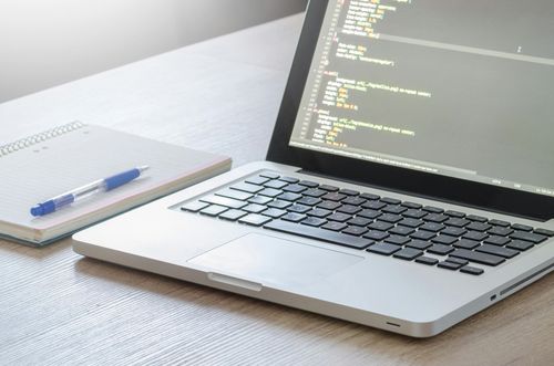 Computer portatile con codice sullo schermo, blocco note e penna su una scrivania di legno.