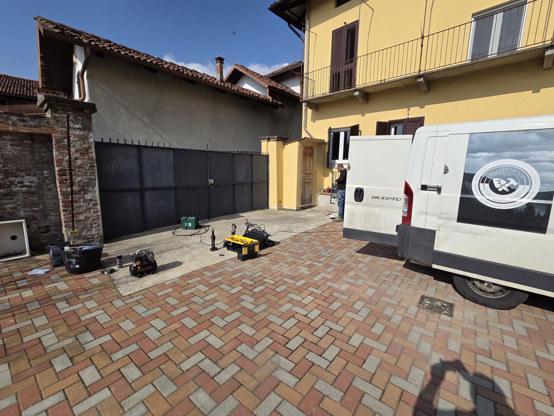 Cortile con pavimentazione in mattoni, attrezzi a terra e un furgone di servizio bianco accanto a un edificio giallo.