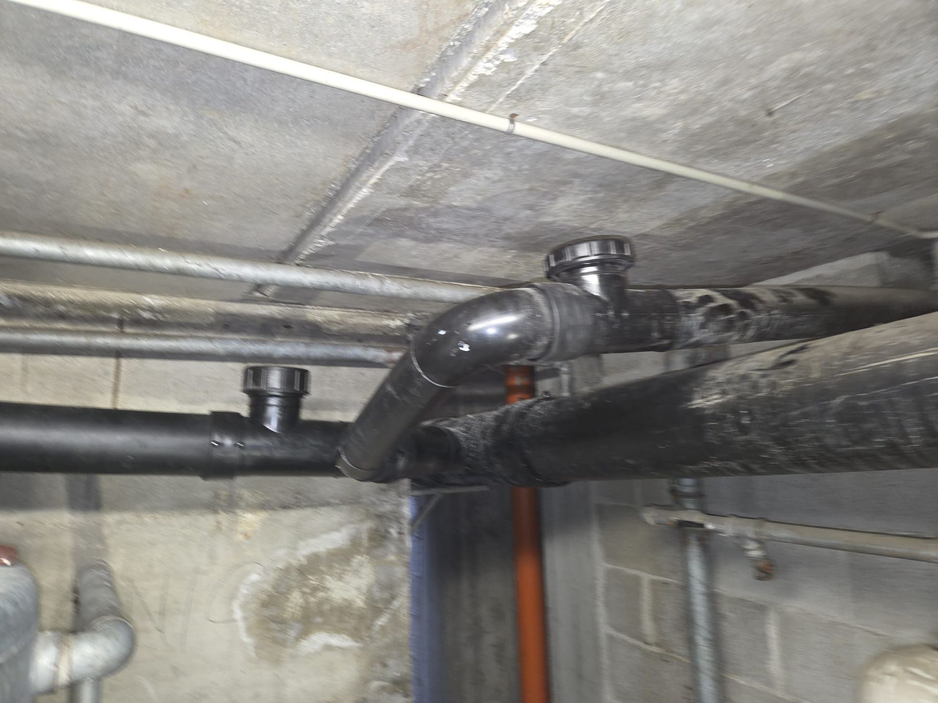 Tubi neri e un raccordo a gomito in PVC passano sotto un soffitto di cemento in uno scantinato o in un vespaio.