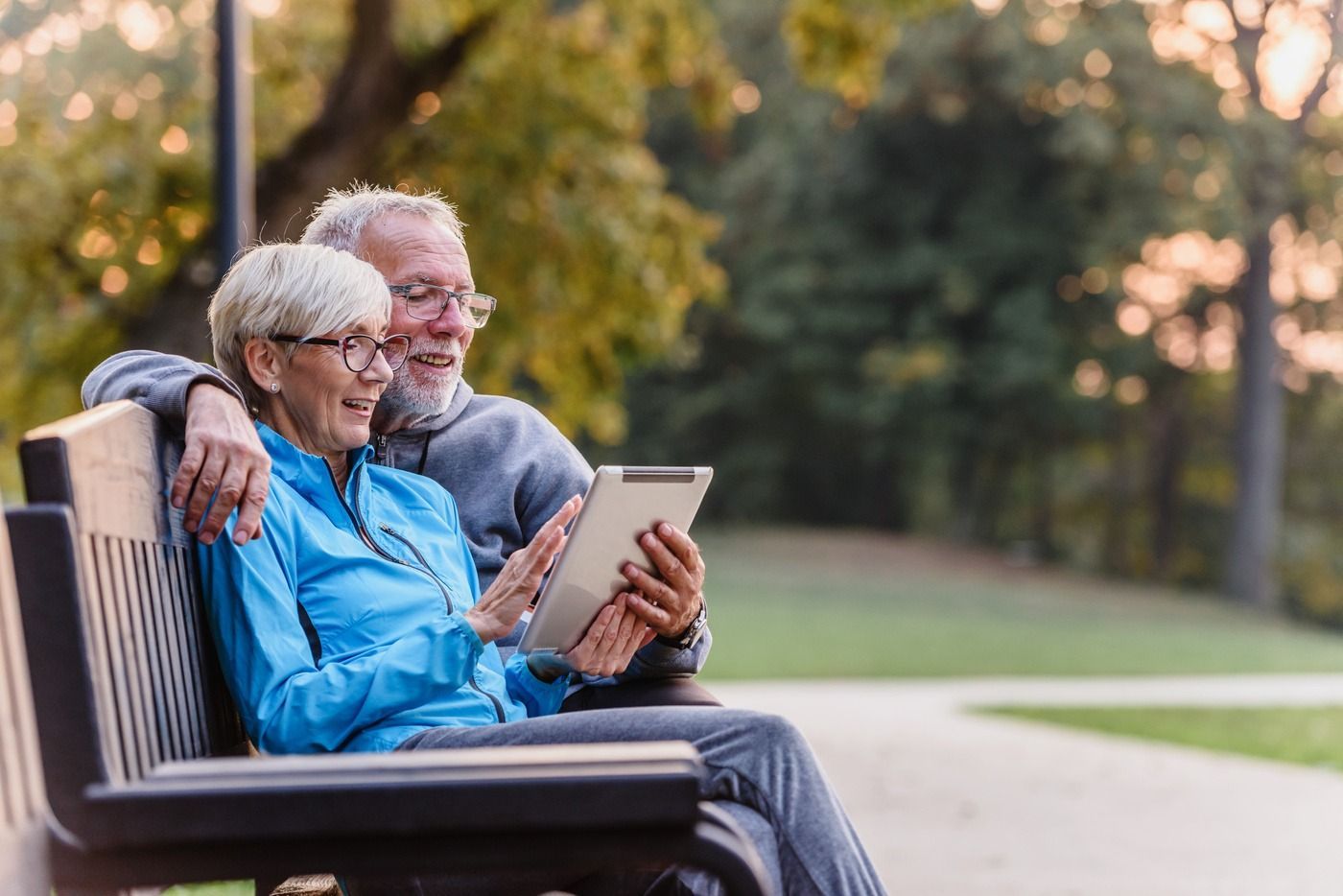 Two people sitting on a park bench, smiling at a tablet in a sunlit park.