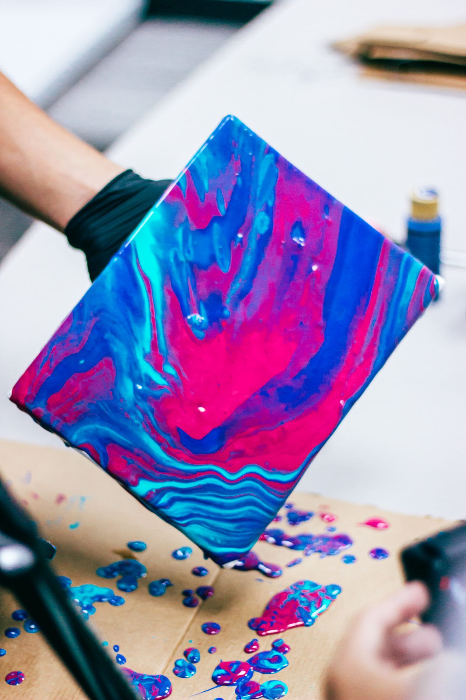 A hand tilting a canvas of hot pink and electric blue poured paint
