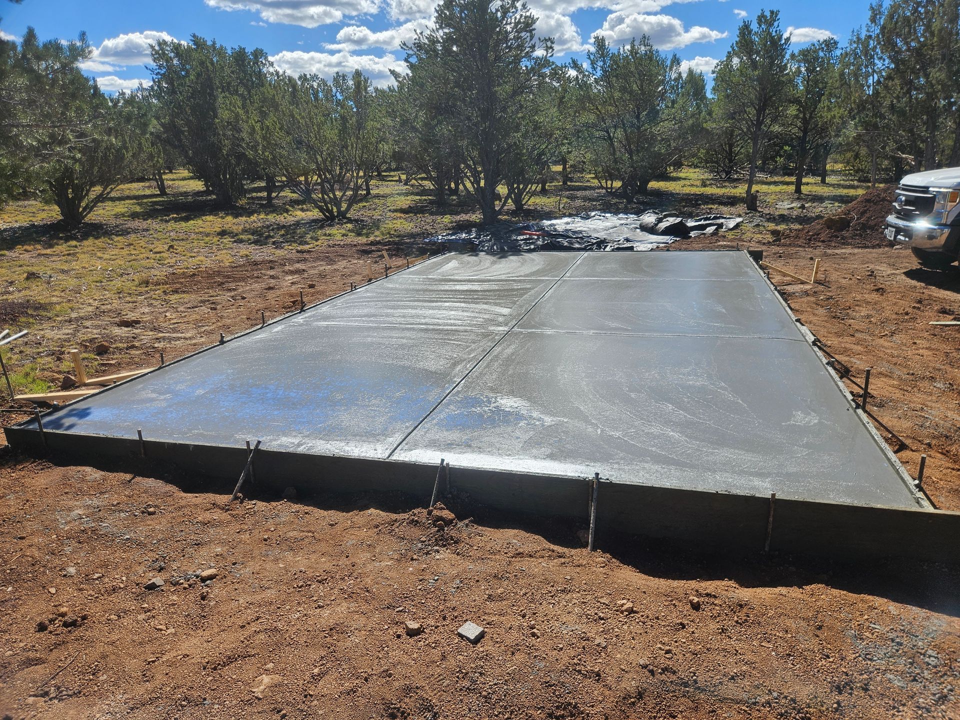 Fresh concrete slab foundation in a rural dirt clearing with trees and a parked vehicle nearby