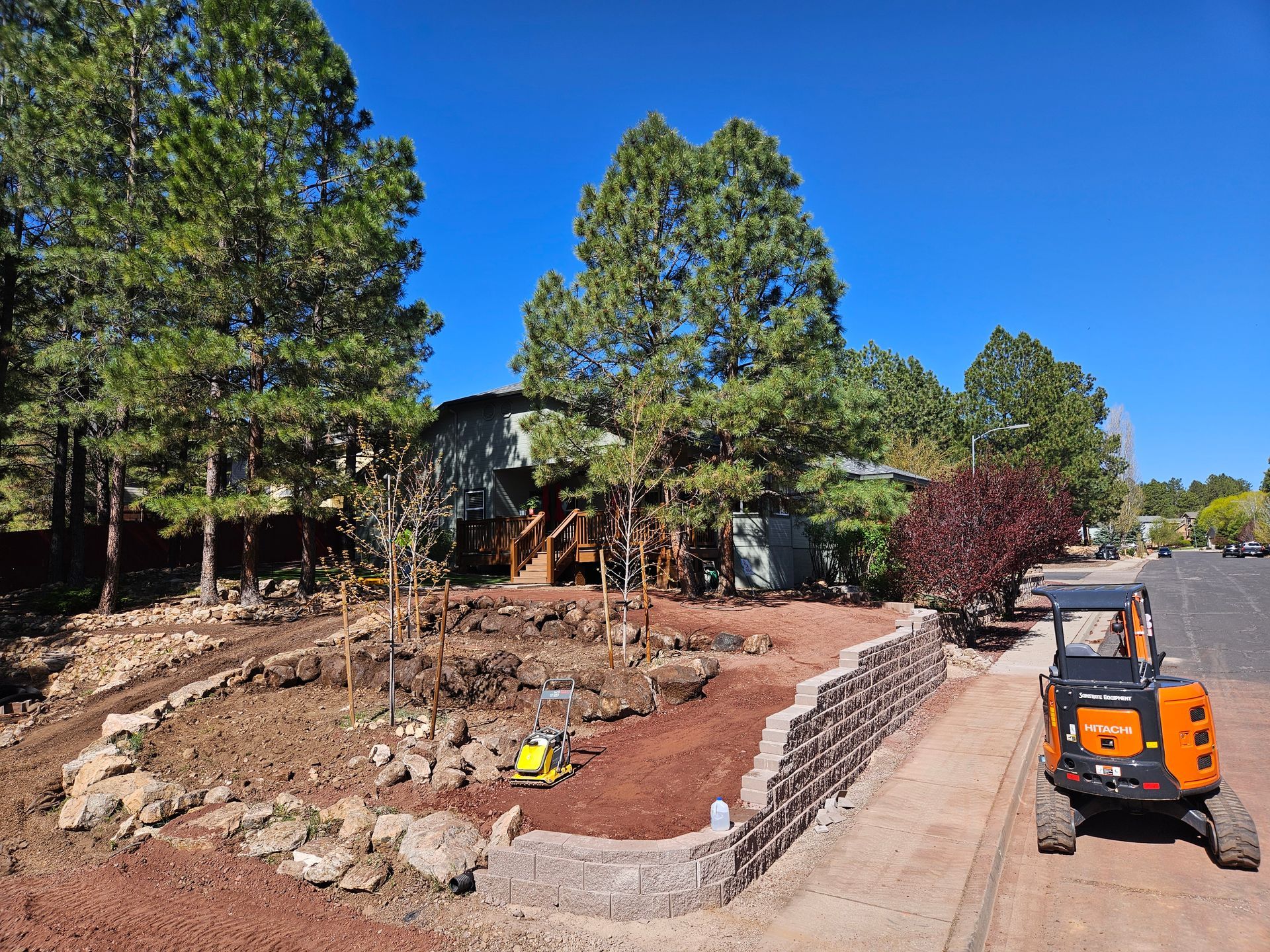 Landscape construction site with stone retaining wall, trees, and an orange skid steer on a paved path
