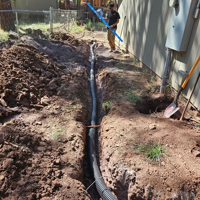 Trench beside a house with black corrugated pipe laid in the ground and a worker in the background
