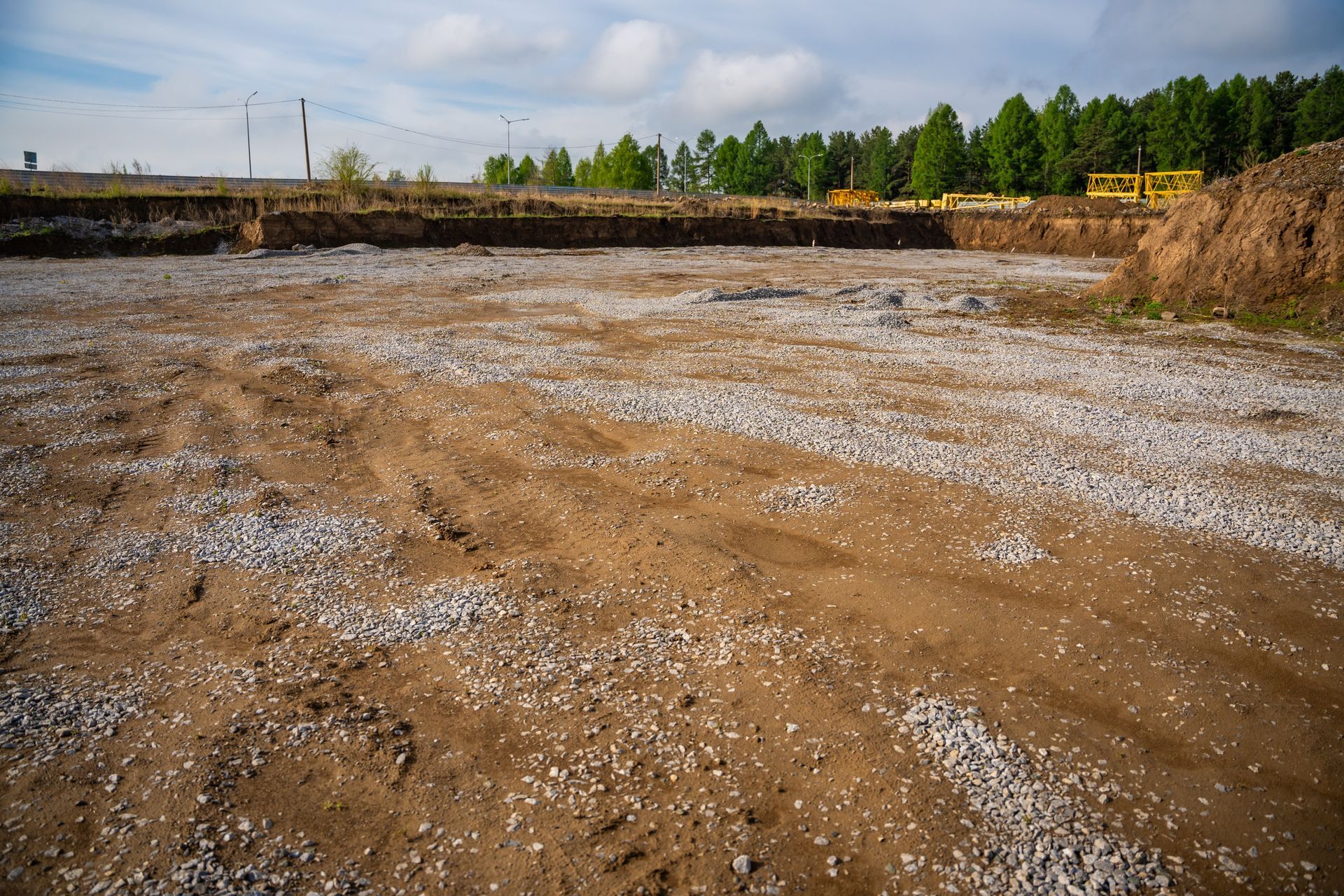 Wide muddy gravel lot with puddles, bordered by trees and a dirt embankment under a cloudy sky