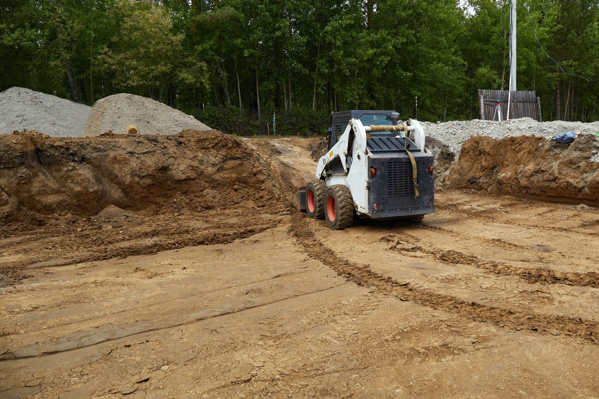 White skid-steer loader working on a dirt construction site with piles of soil and trees in the background
