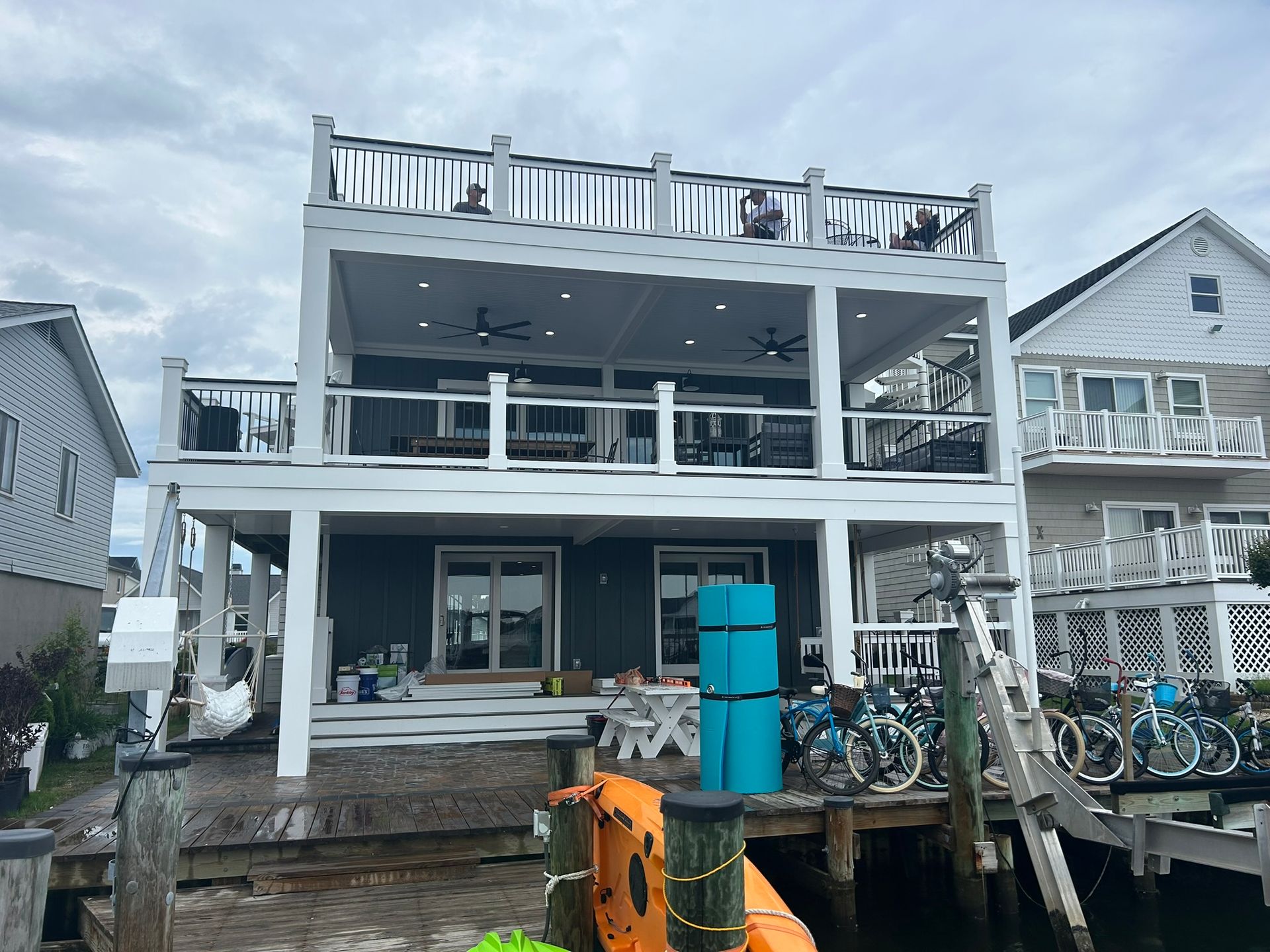 Three-story house with decks on a waterfront. White railings, teal siding, bikes on dock. Overcast sky.