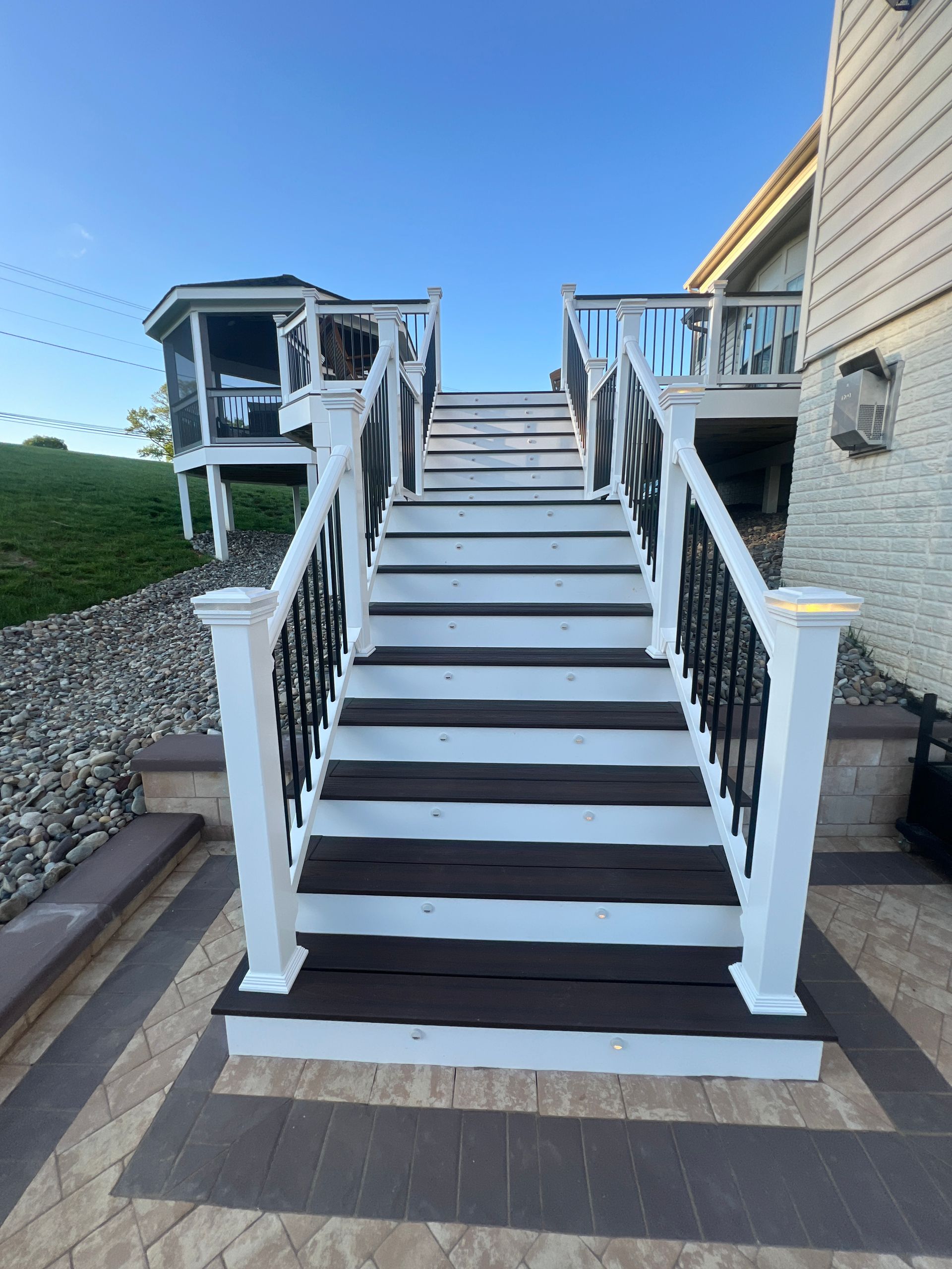 Wooden staircase with white posts and black railings leads up to a deck with a gazebo, clear blue sky overhead.