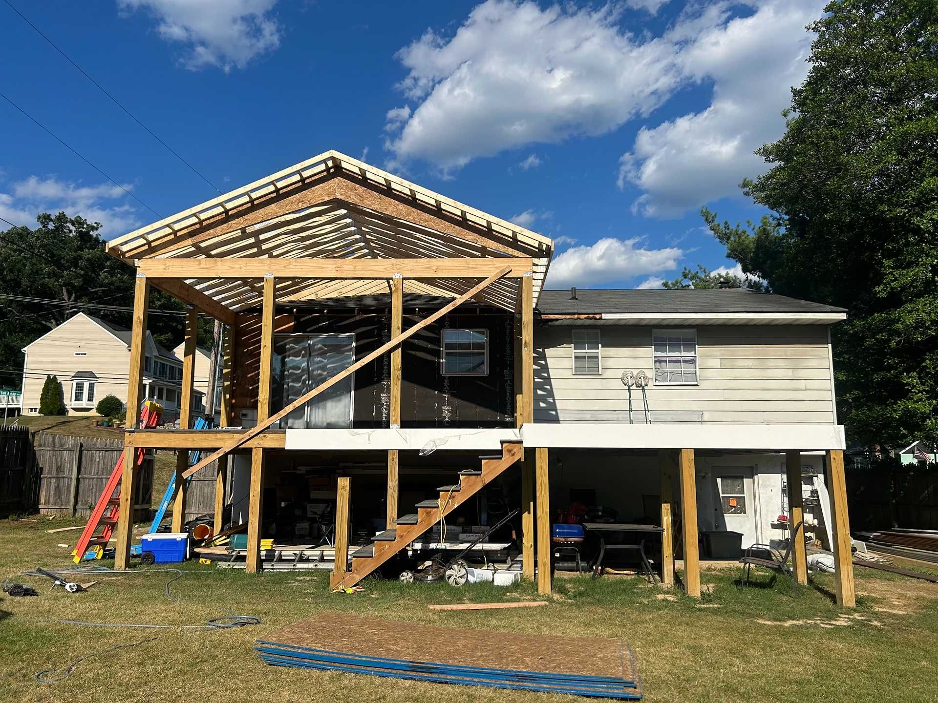 Construction of a raised deck and roof addition on a house. Wooden framework, stairs, and blue sky.