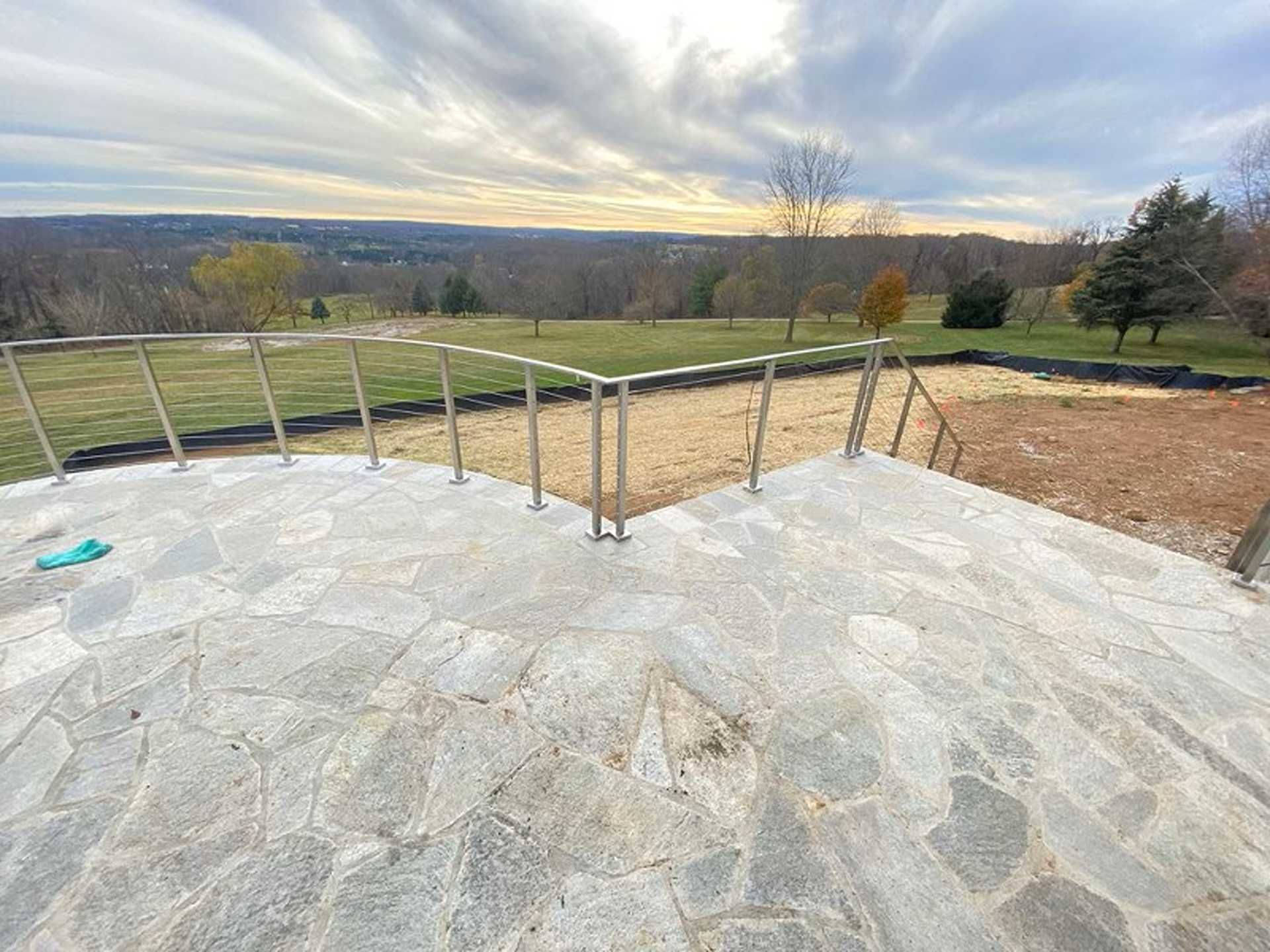 Stone patio with stainless steel railing overlooking a field and trees under a cloudy sky.