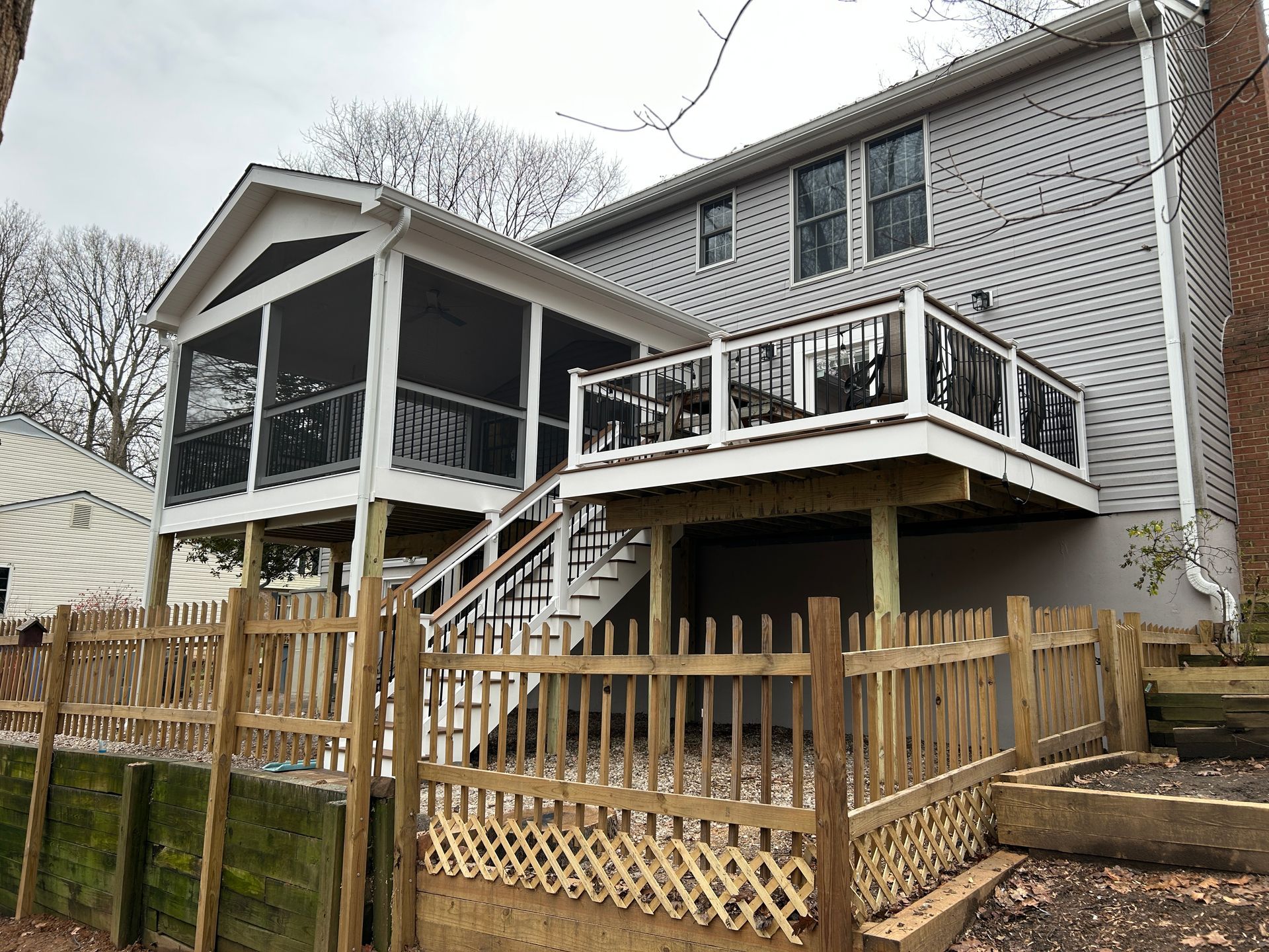 Back of a two-story house with a screened porch and deck. Wooden fence encloses a lower patio area.