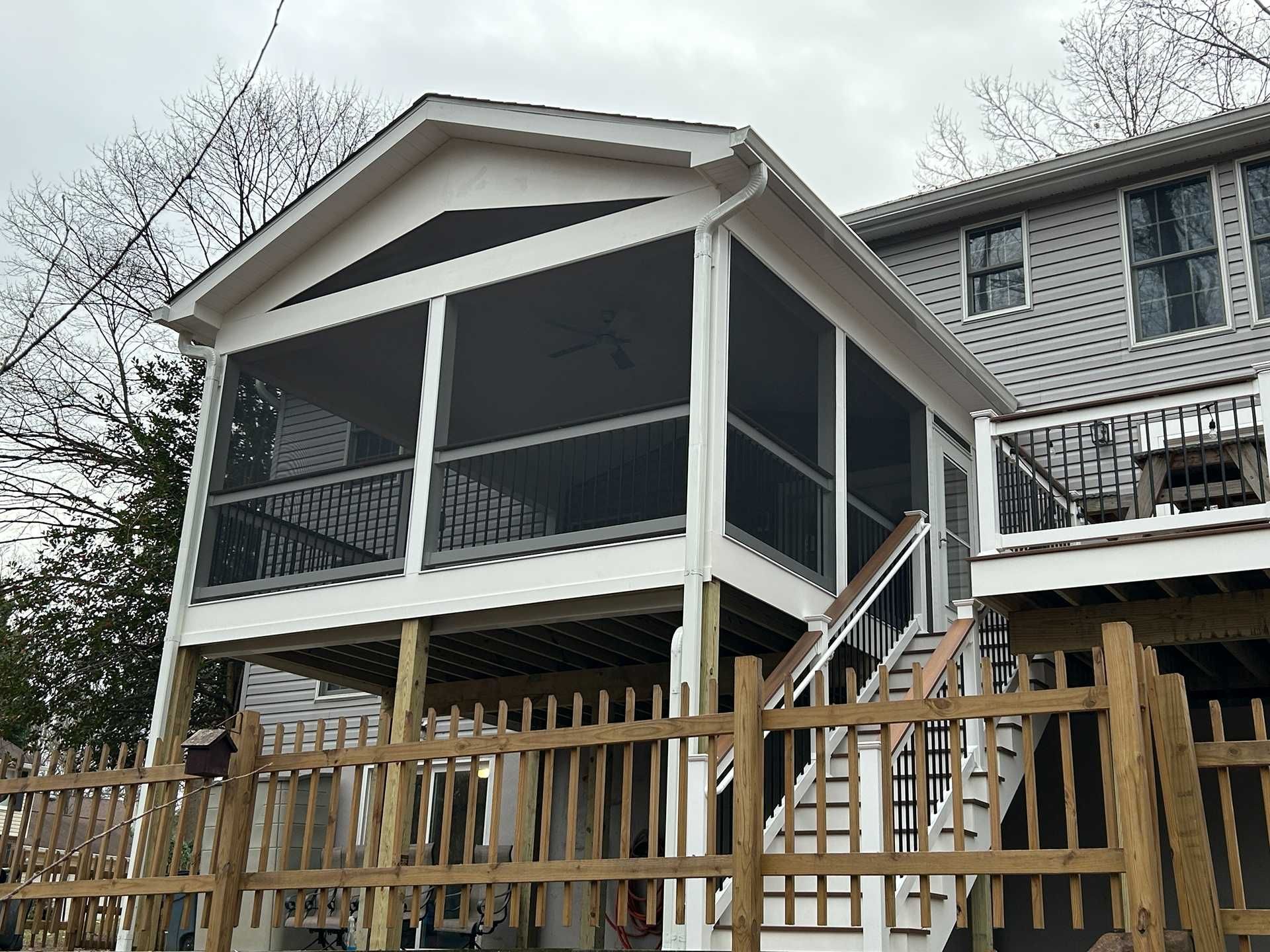 Screened porch with stairs attached to a house with gray siding; a wooden fence in the foreground.