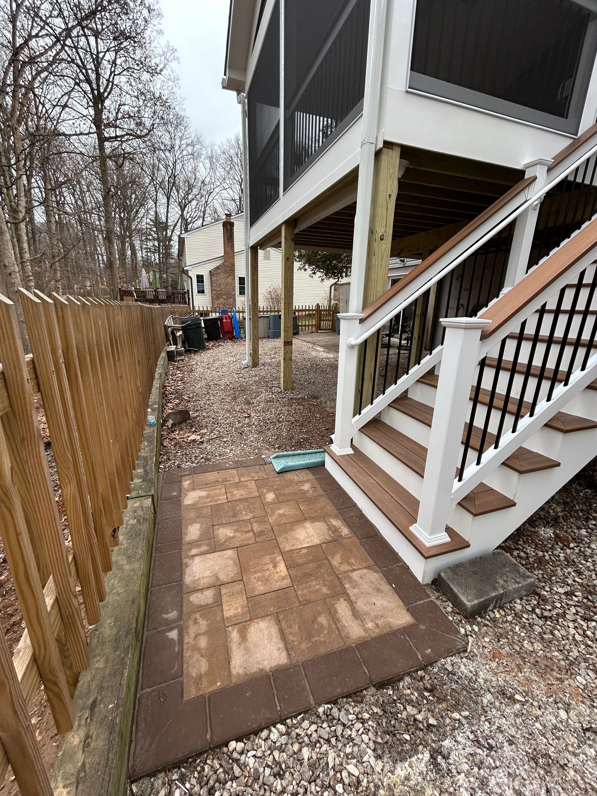 Patio with brick pavers, wooden fence, and deck stairs. Brown and beige tones.
