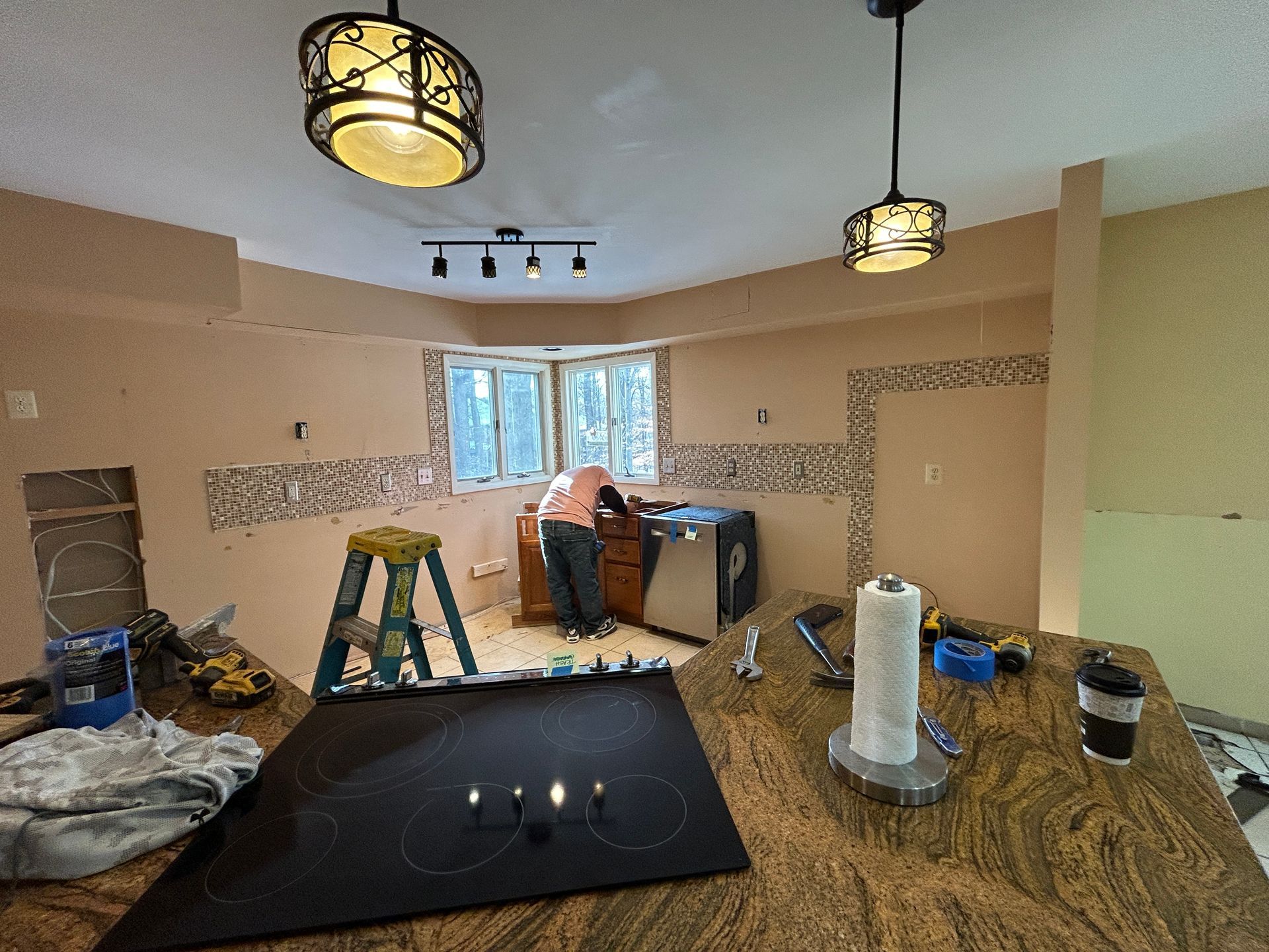 Kitchen renovation in progress: person working near a window, countertops, cabinets, and various tools.