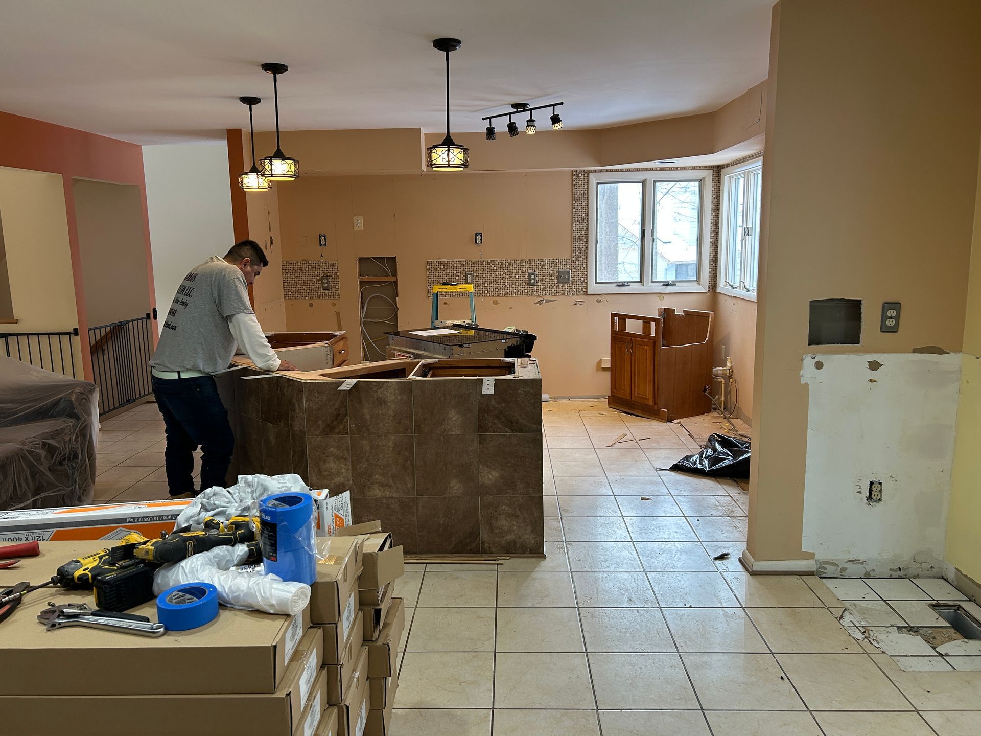Man working on kitchen renovation, removing flooring and cabinets. Beige walls, open layout.