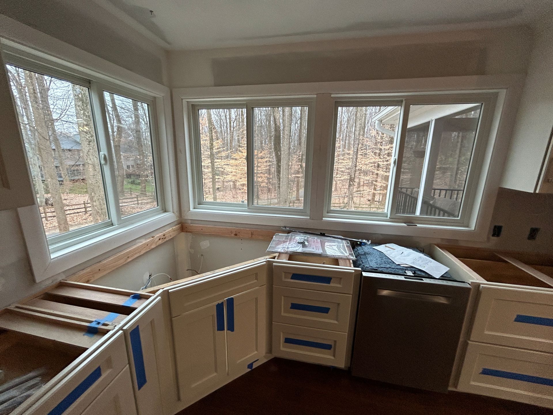 Kitchen renovation in progress: white cabinets installed, dishwasher visible, windows overlooking trees.