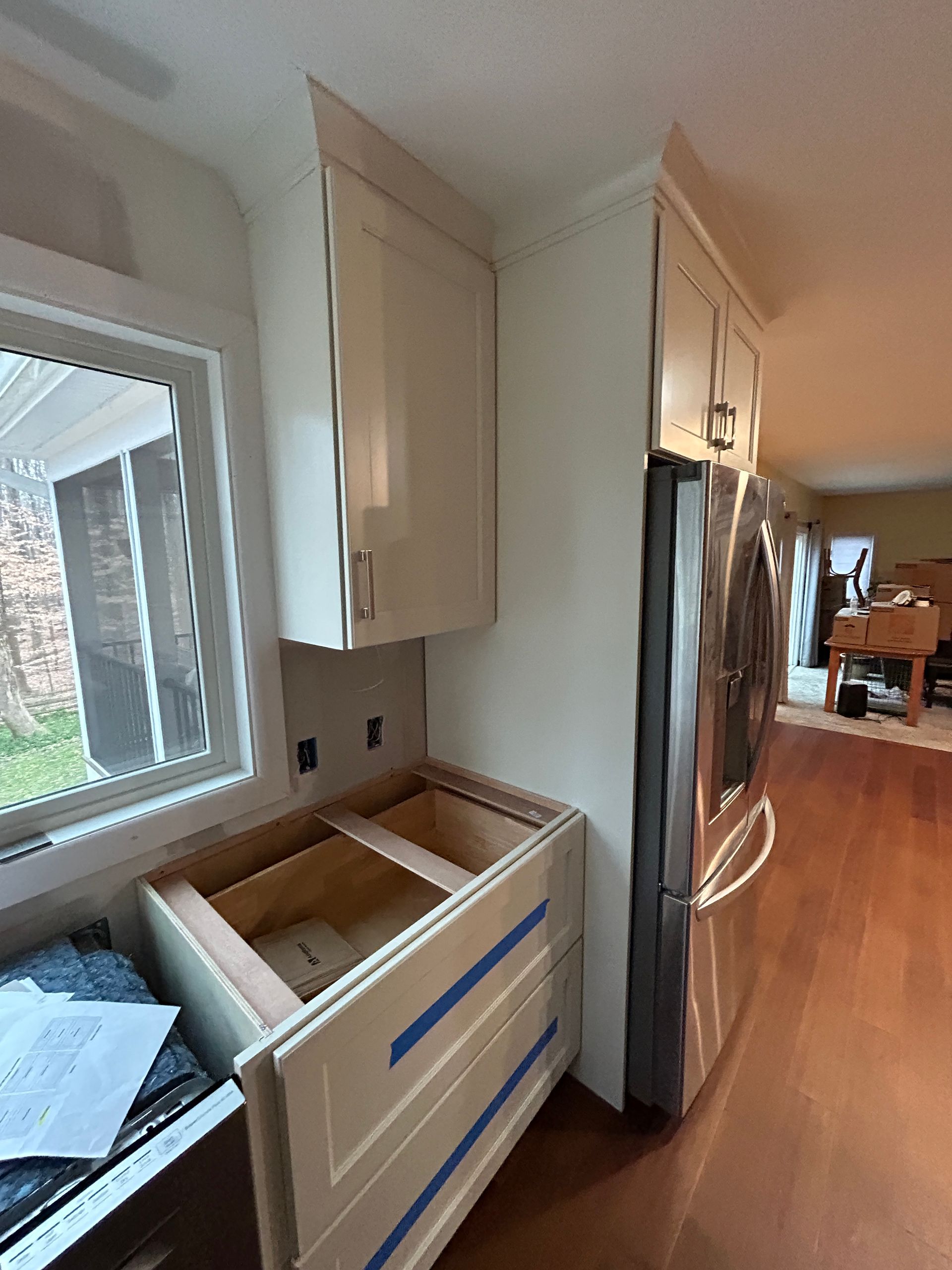 Kitchen cabinets being installed near a window and refrigerator. Unfinished lower cabinet with open door.
