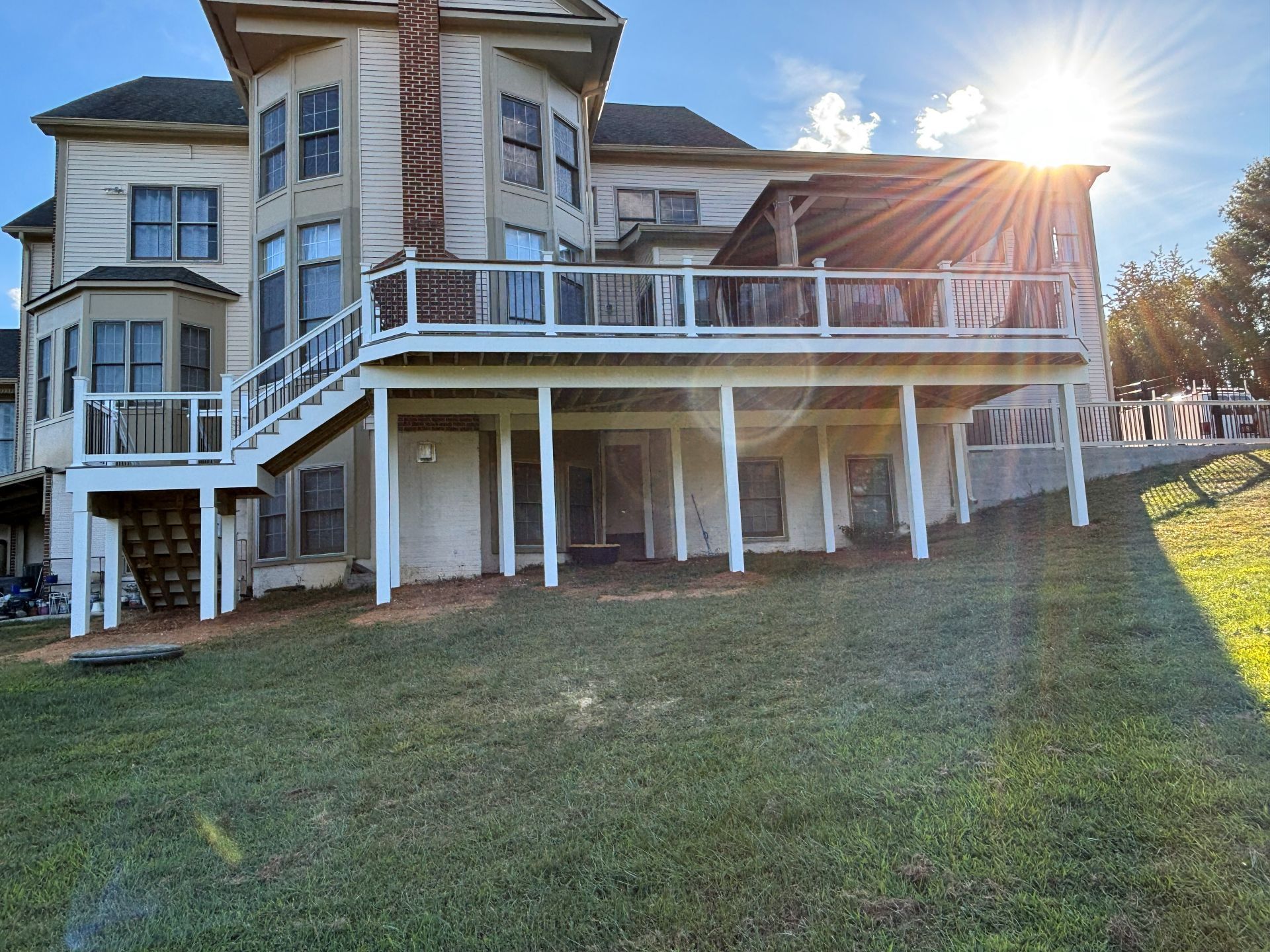 Large house with a multi-level deck, set on a grassy hill; sun shining brightly in the sky.