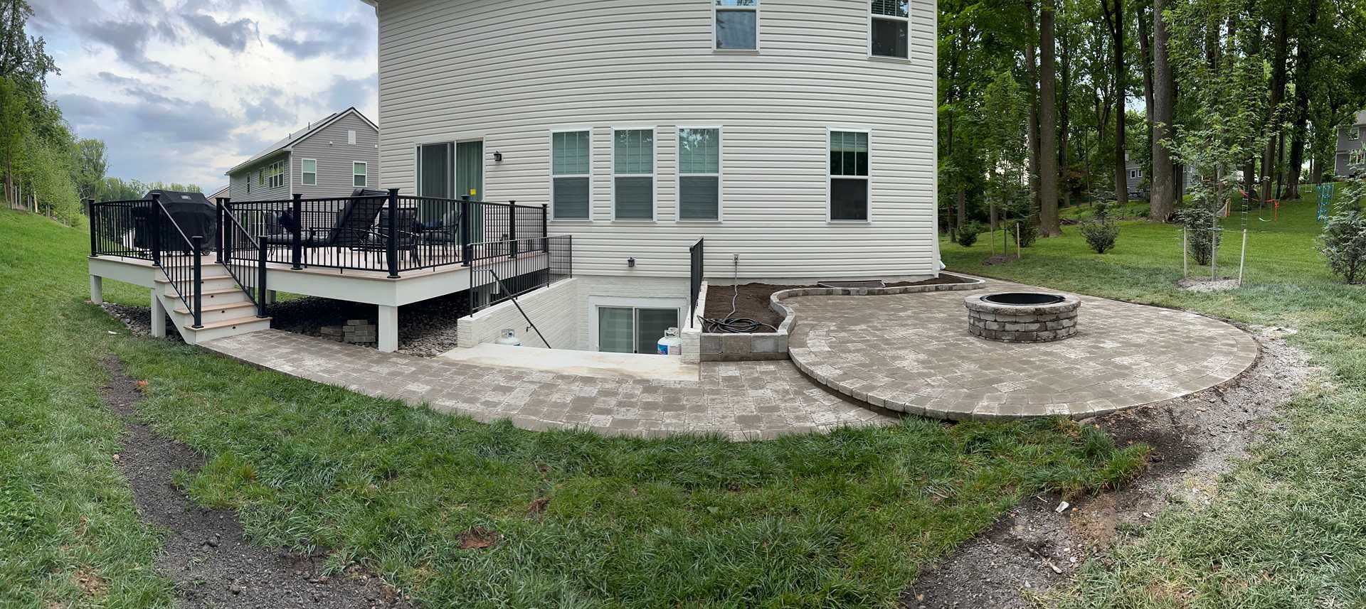 Back of a two-story house with a deck, patio with fire pit, and a grassy yard with trees.