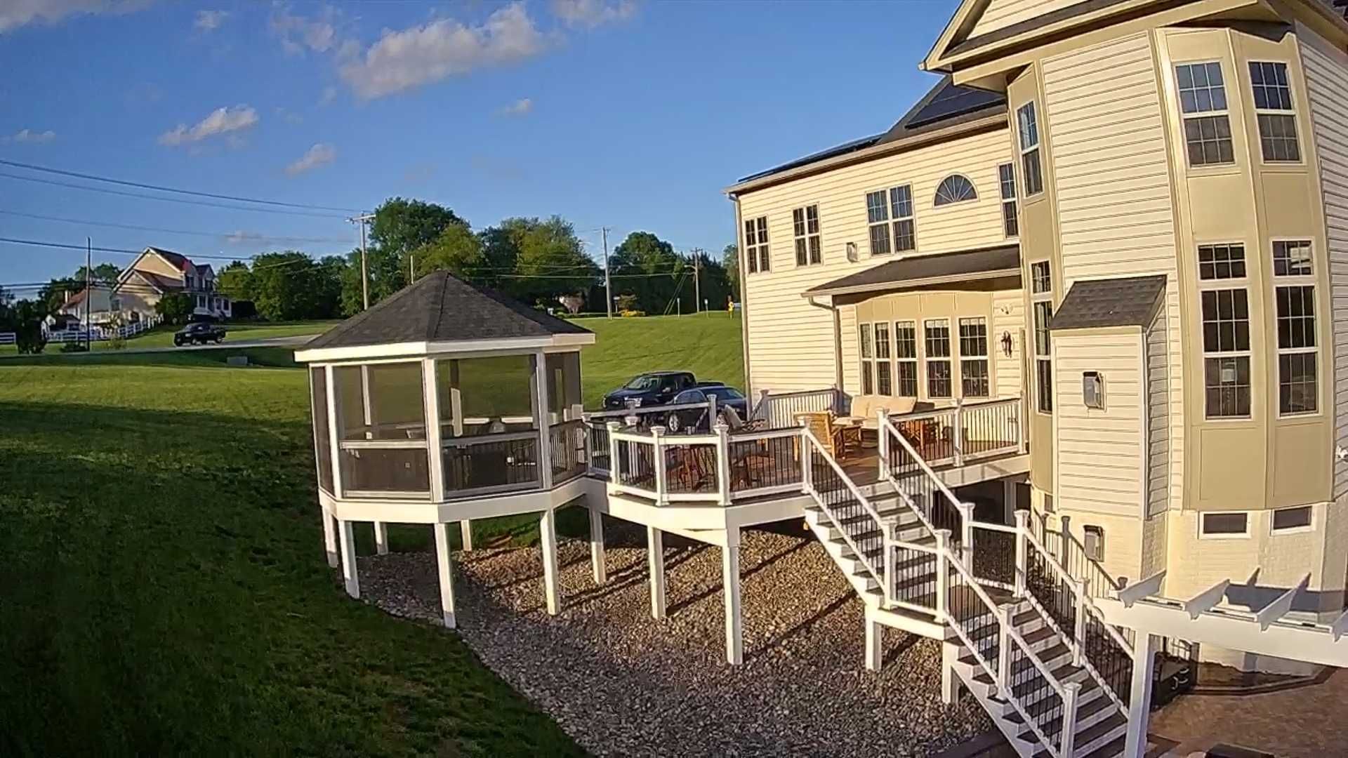 A two-story house with a wooden deck and gazebo in a grassy yard under a blue sky.