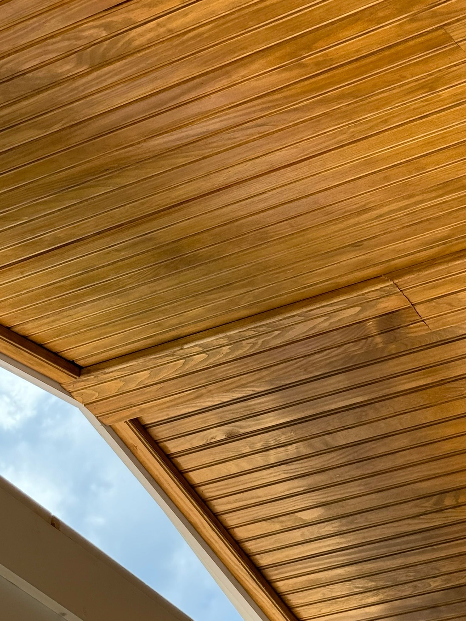 Wood-paneled ceiling with rows of grooved slats, seen from below, with a glimpse of blue sky.