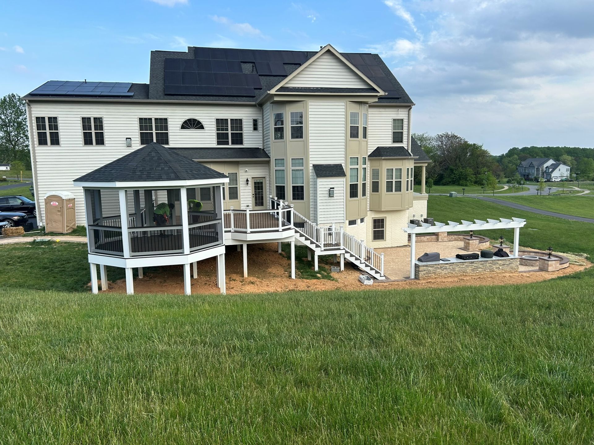 Back view of a two-story beige house with a gazebo, deck, and outdoor kitchen. Solar panels are on the roof.