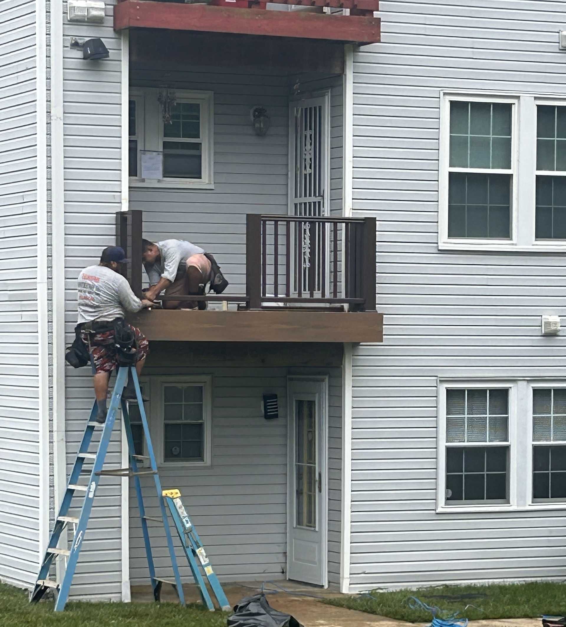 Two workers installing a deck on an apartment building. One on a ladder, one on the deck.