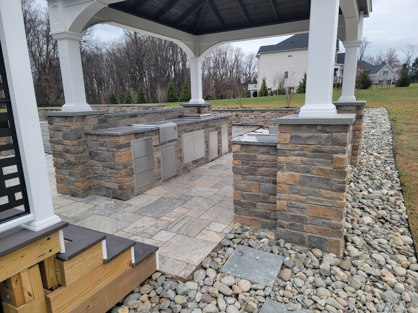 Outdoor kitchen under a gazebo. Stone facade with grilling area, paved patio, and rock border.
