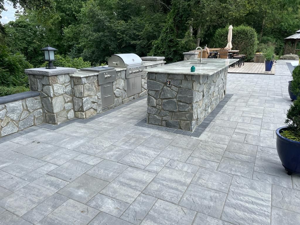 Stone outdoor kitchen with grill and counter on a stone patio, surrounded by trees.
