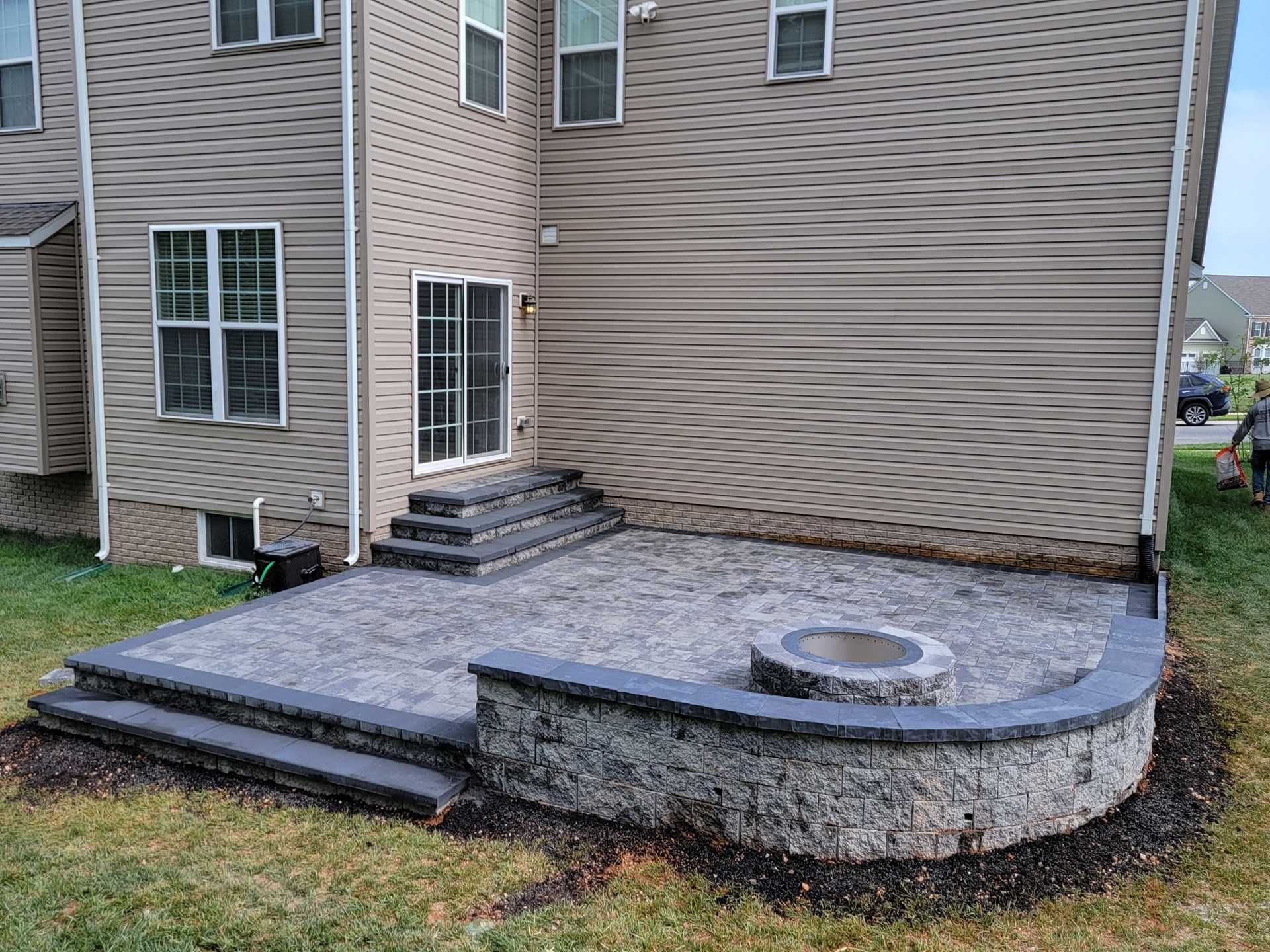 A paver patio with steps and a low retaining wall around a fire pit, adjacent to a house.