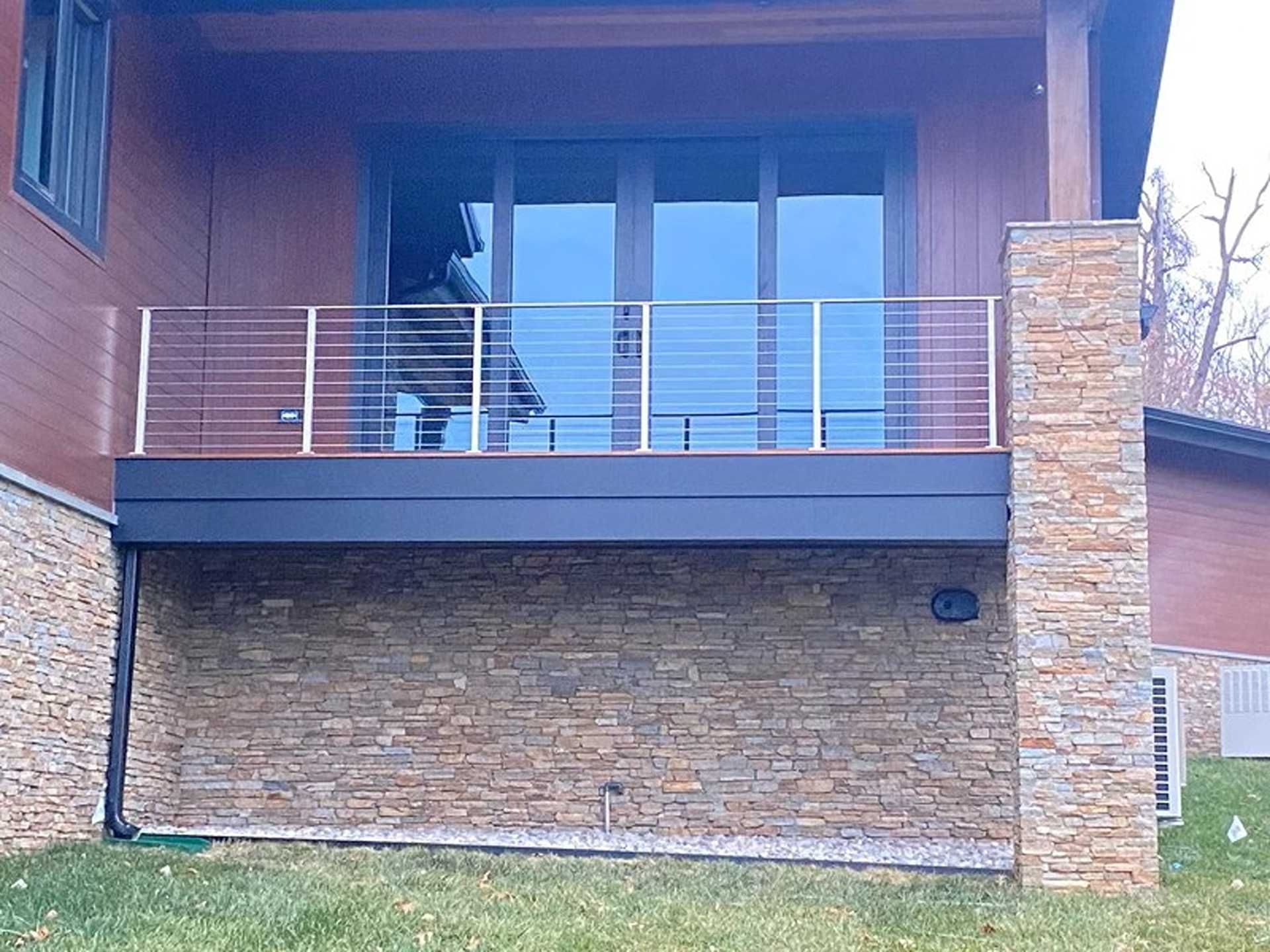 Deck with cable railings, stone facade below. Large windows reflect a person.