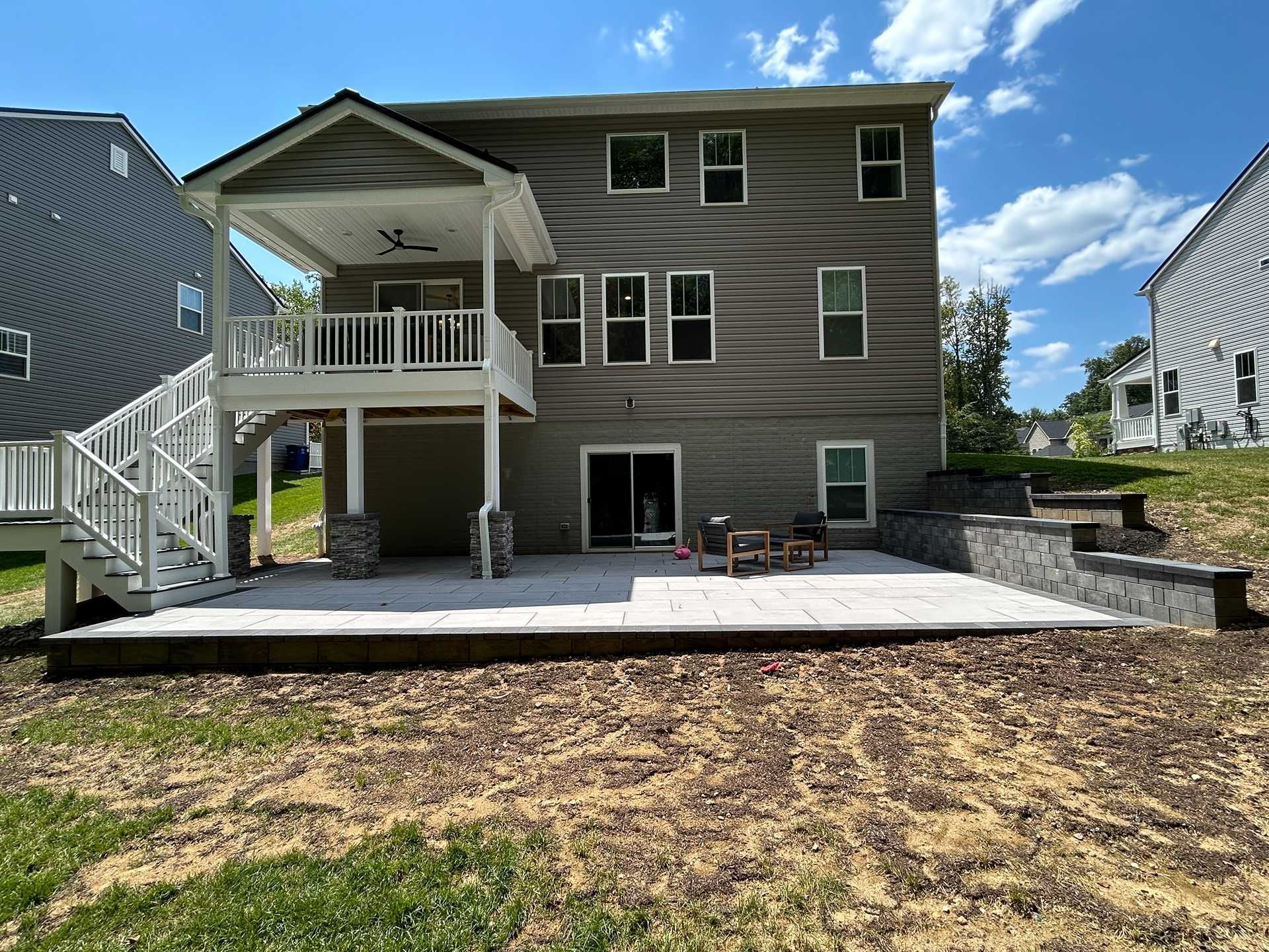 Backyard patio with two-story house, a deck, and retaining walls under a blue sky.