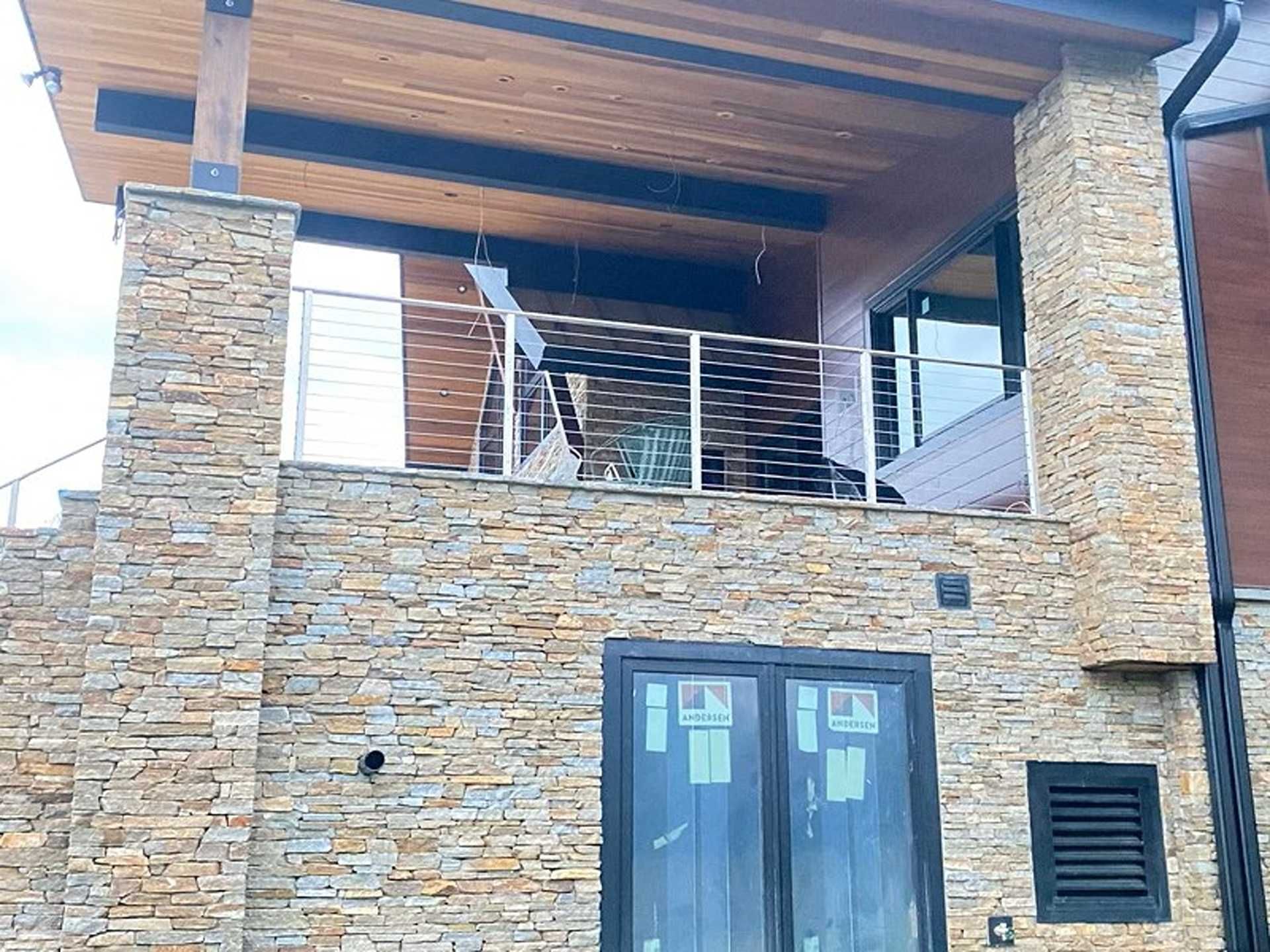 Stone-clad building with a balcony featuring stainless steel railings. Wooden ceiling and a dark window frame.