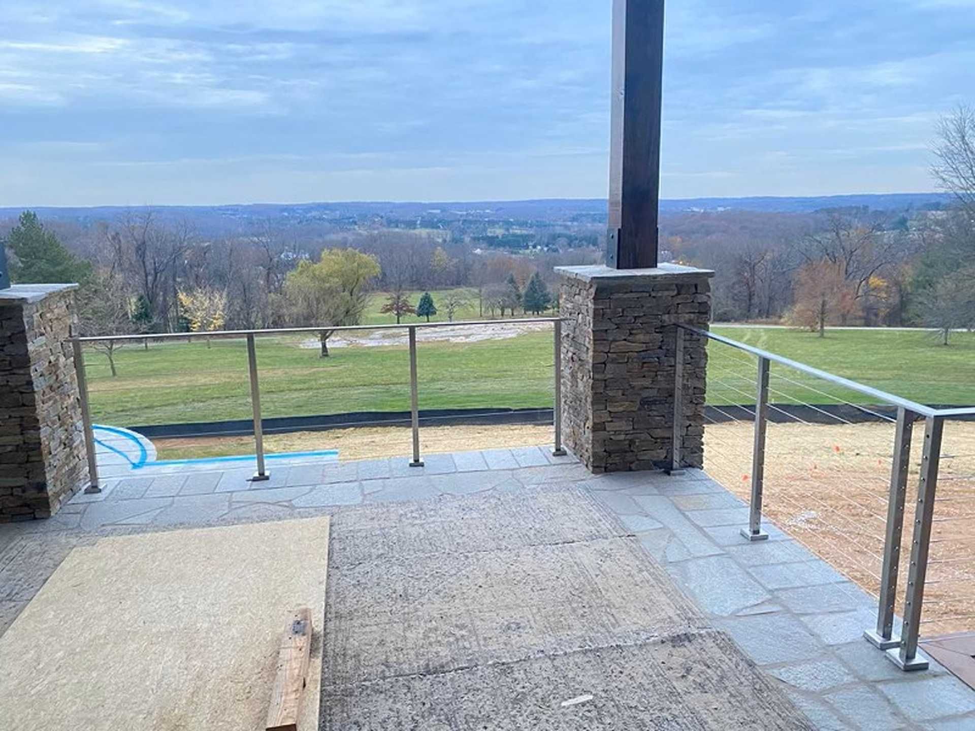 Patio with stone pillars and railing, overlooking a green field and trees on a cloudy day.