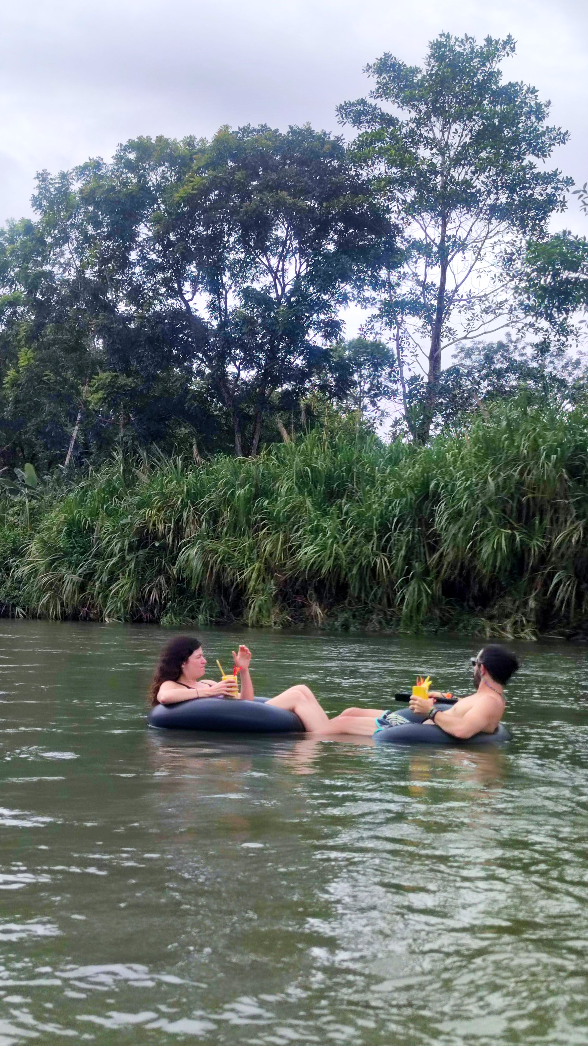 Two people floating on inflatable rings in a calm river, with tall grass and trees on the bank.