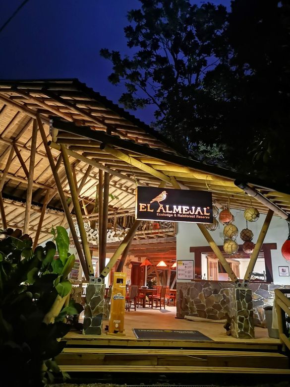 Open-air restaurant entrance at dusk with warm lights, wooden beams, and greenery