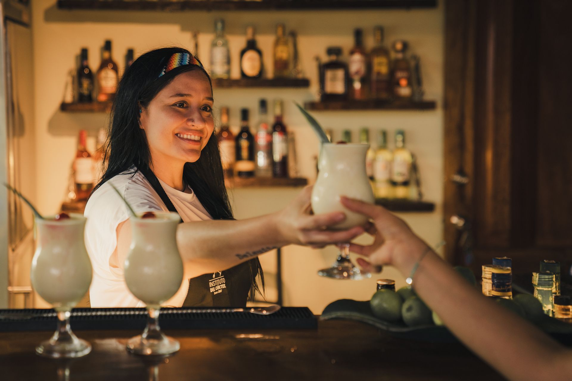 Bartender handing a drink across a bar, with cocktails and bottles softly lit in the background.
