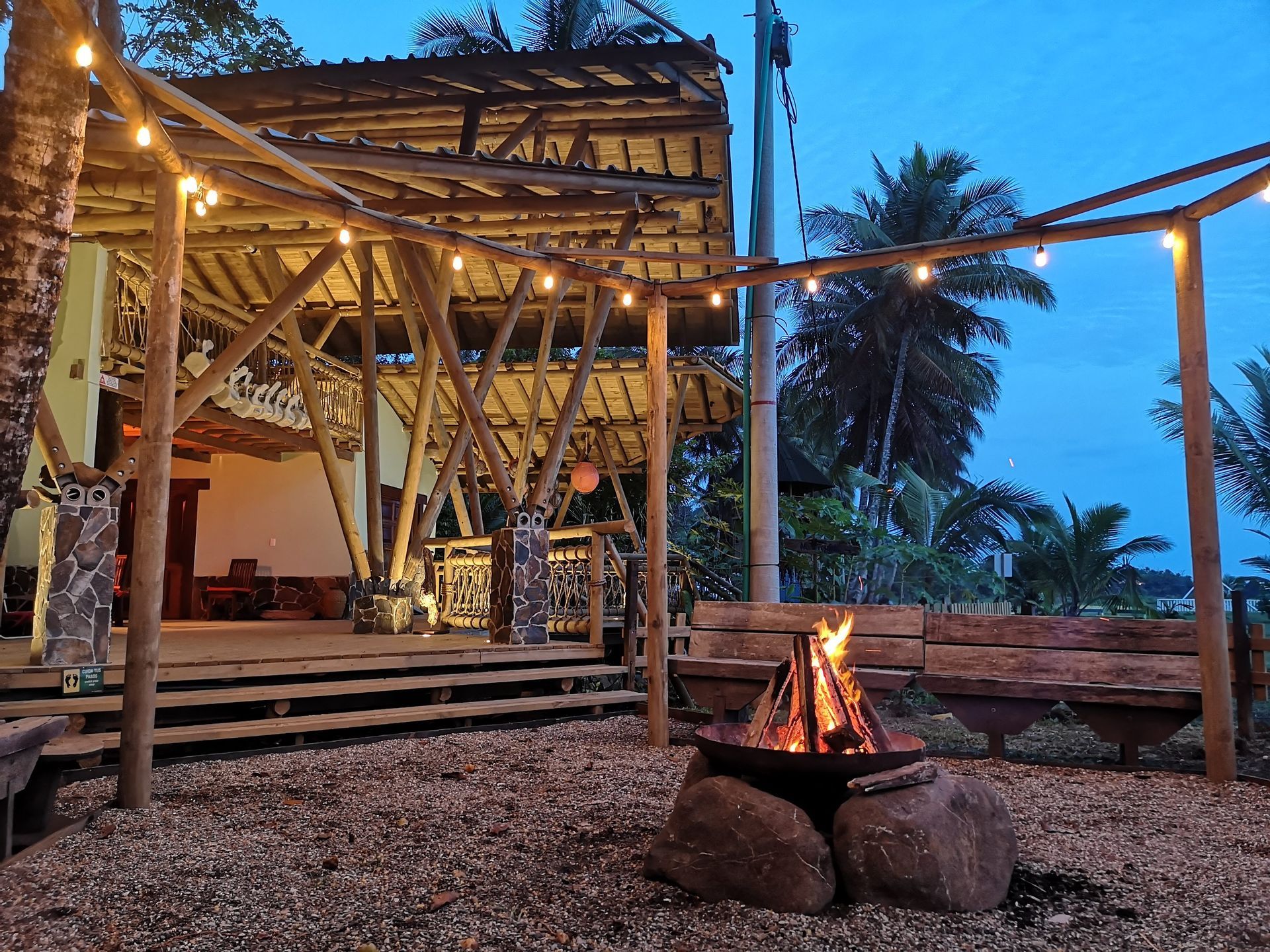 Outdoor fire pit beside a bamboo hut at dusk, with string lights and a palm tree in the background