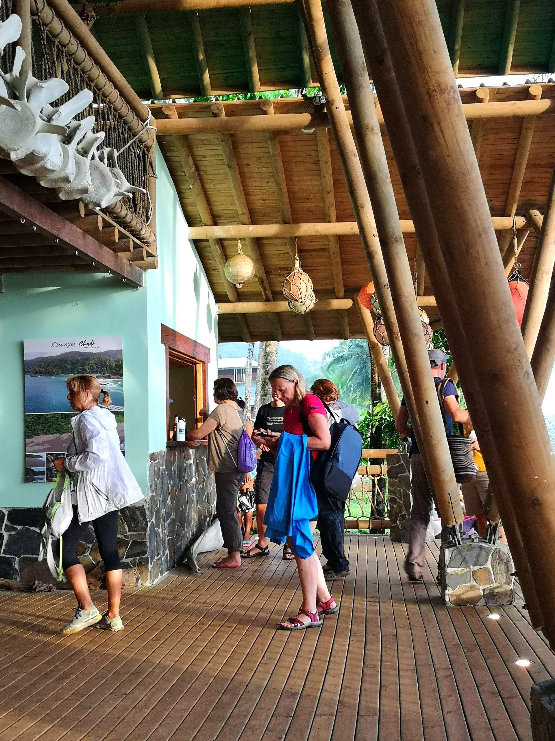 People standing in a bamboo-roofed open-air shelter on a wooden deck, with tables and railings nearby.