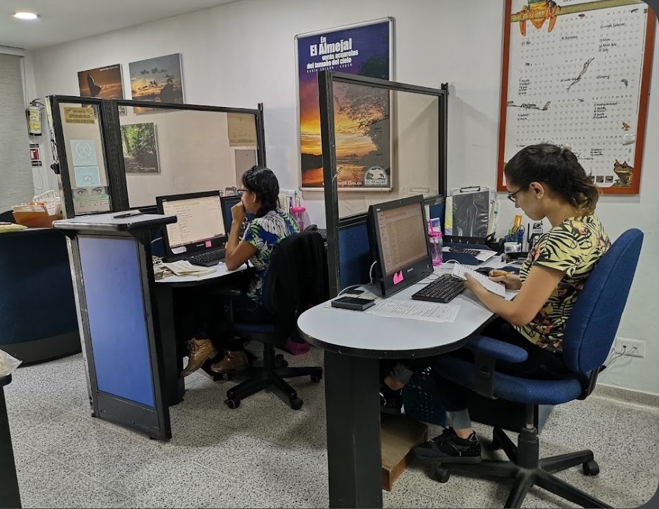 Two people working at desks in a small office with blue chairs and computer monitors