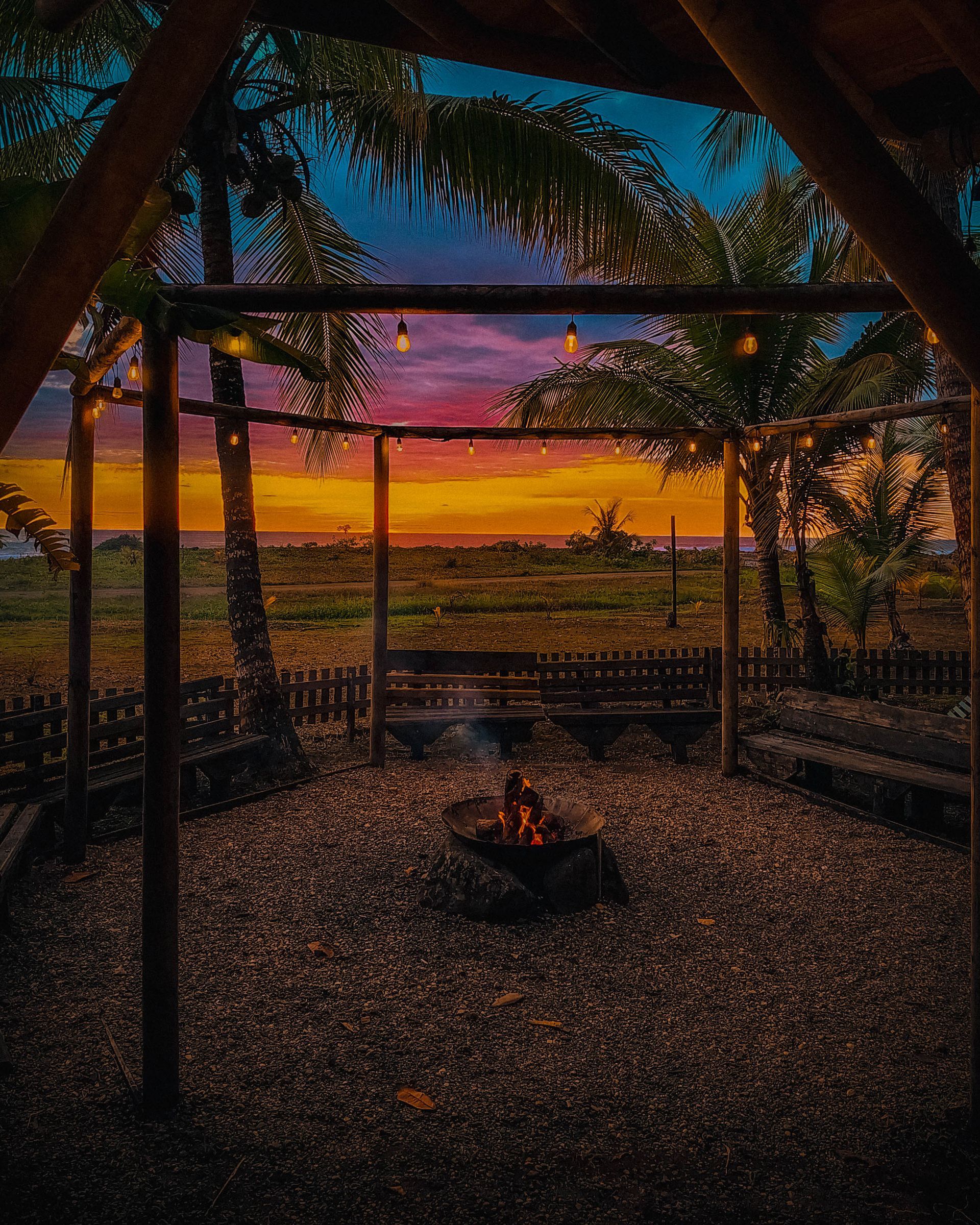 Tropical gazebo at sunset, with a small campfire and colorful sky over a distant field.