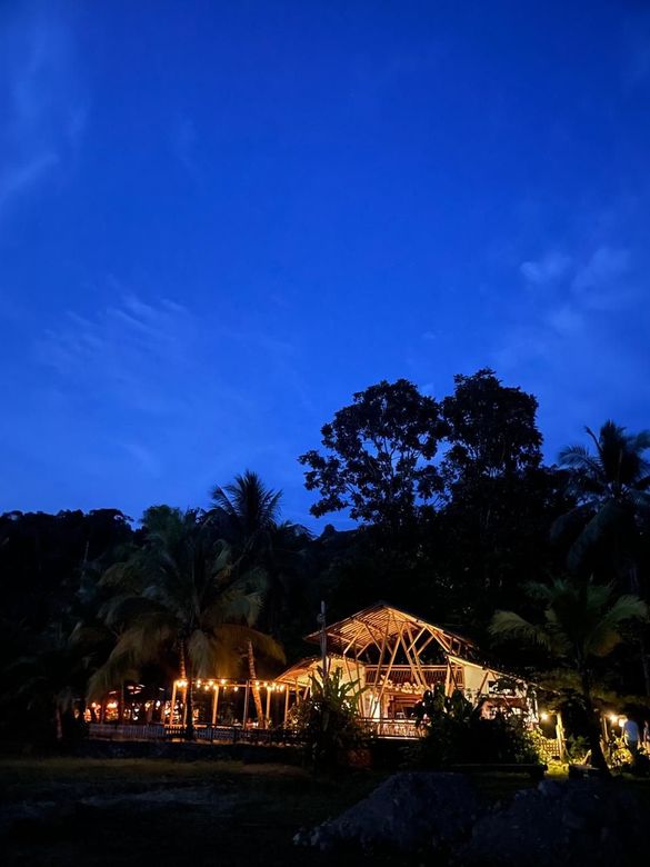 Tropical resort lit at dusk beneath a deep blue sky, with palm trees and thatched-roof buildings.