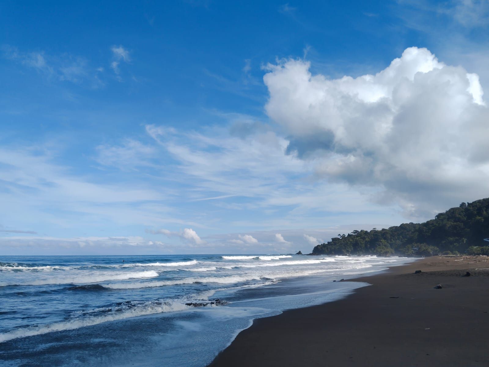 Sunny beach with waves, dark sand, and a large white cloud over the ocean