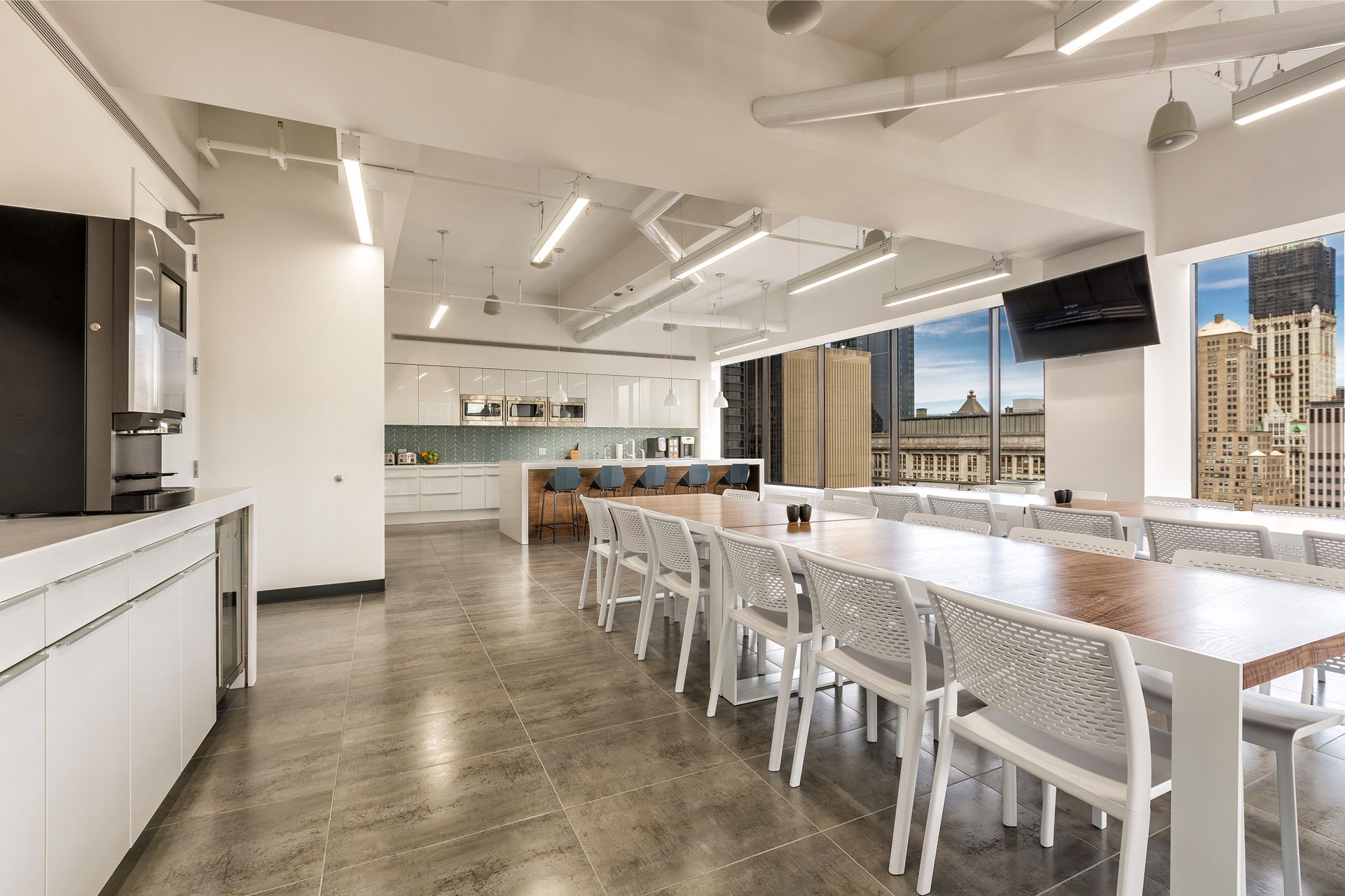 Modern office break room with long table and city view. White and grey decor, kitchen in background.
